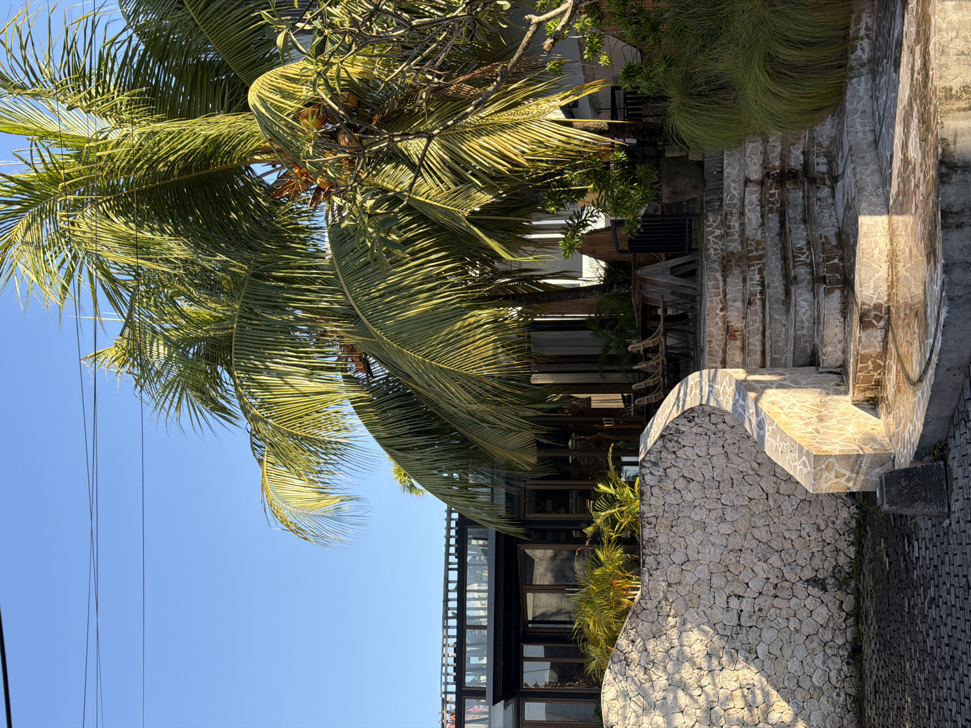 The image shows a sunlit exterior scene in North Kuta, Indonesia, featuring a building with dark-framed windows and a balcony, surrounded by lush tropical vegetation. In the foreground, a curved stone wall leads to a set of outdoor stairs. Palm trees with coconuts are prominent, with their fronds casting shadows. The ground is paved with dark interlocking bricks, and there are some planters with greenery. The overall impression is of a tropical restaurant or resort.