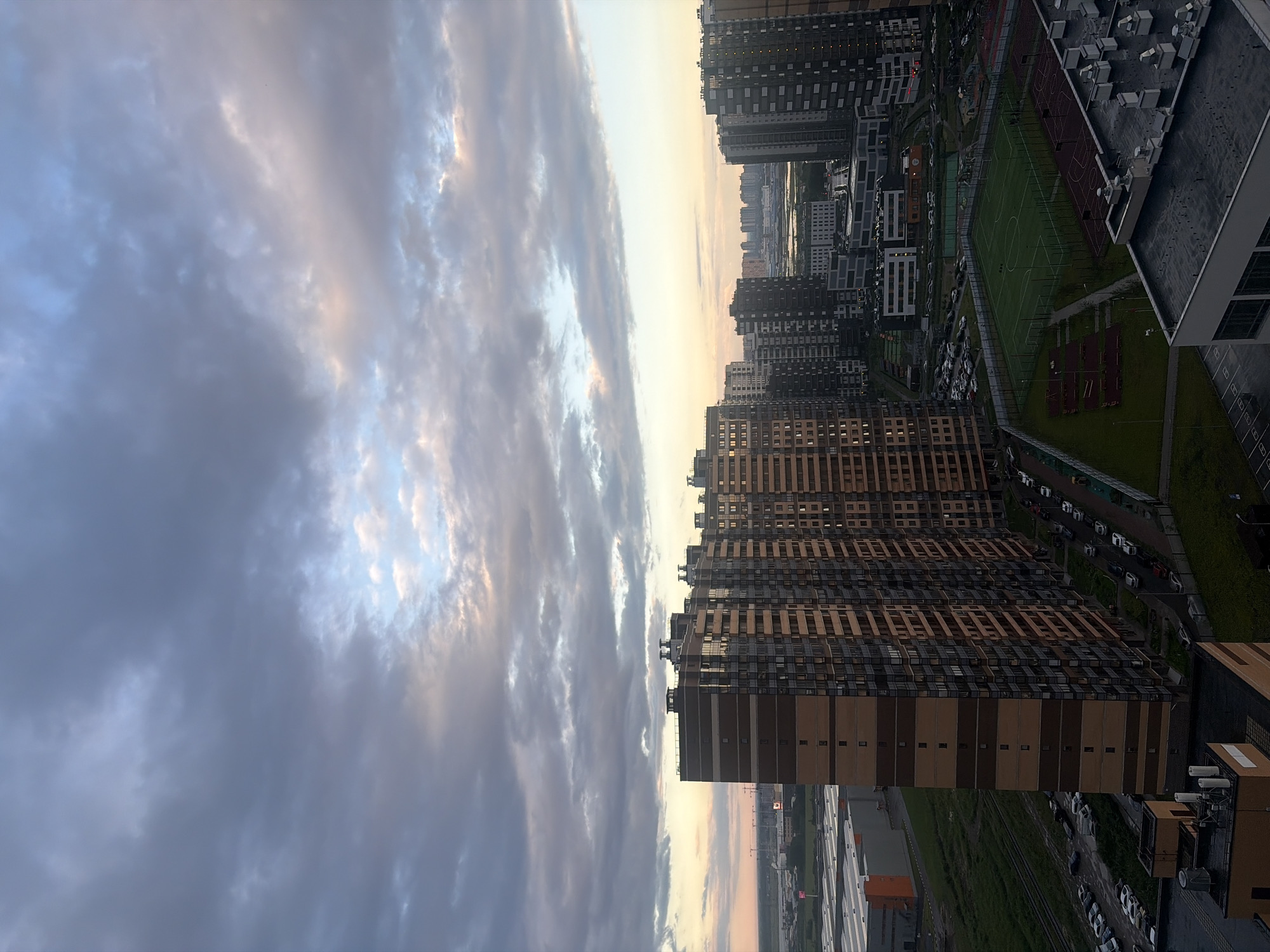 The image depicts an aerial view of a residential area in Saint Petersburg, Russia, during what appears to be the late afternoon or early evening. The sky is filled with dramatic, overcast clouds tinged with the warm hues of a setting sun, with patches of lighter blue sky visible.
Dominating the foreground are several tall, modern apartment buildings with distinctive brick and glass facades. The building on the left is particularly prominent, featuring horizontal bands of brown and beige brickwork. To its right, a cluster of other high-rise buildings forms a dense urban landscape, with some buildings displaying darker, more contemporary designs.
In the mid-ground, a green, fenced-in sports field, possibly a soccer pitch, is visible, along with other recreational areas like basketball courts. Surrounding the buildings and recreational spaces are parking lots filled with numerous cars, indicating a populated residential area. A railway line runs alongside the sports field, suggesting the presence of transport infrastructure.
In the background, a hazy skyline of further buildings stretches towards the horizon, characteristic of a large city. In the far distance to the left, industrial buildings and construction cranes are visible, hinting at ongoing development or industrial zones within the city. The overall scene is one of a contemporary urban environment with residential high-rises and associated amenities under a dynamic, cloudy sky. There are no visible people in the image.