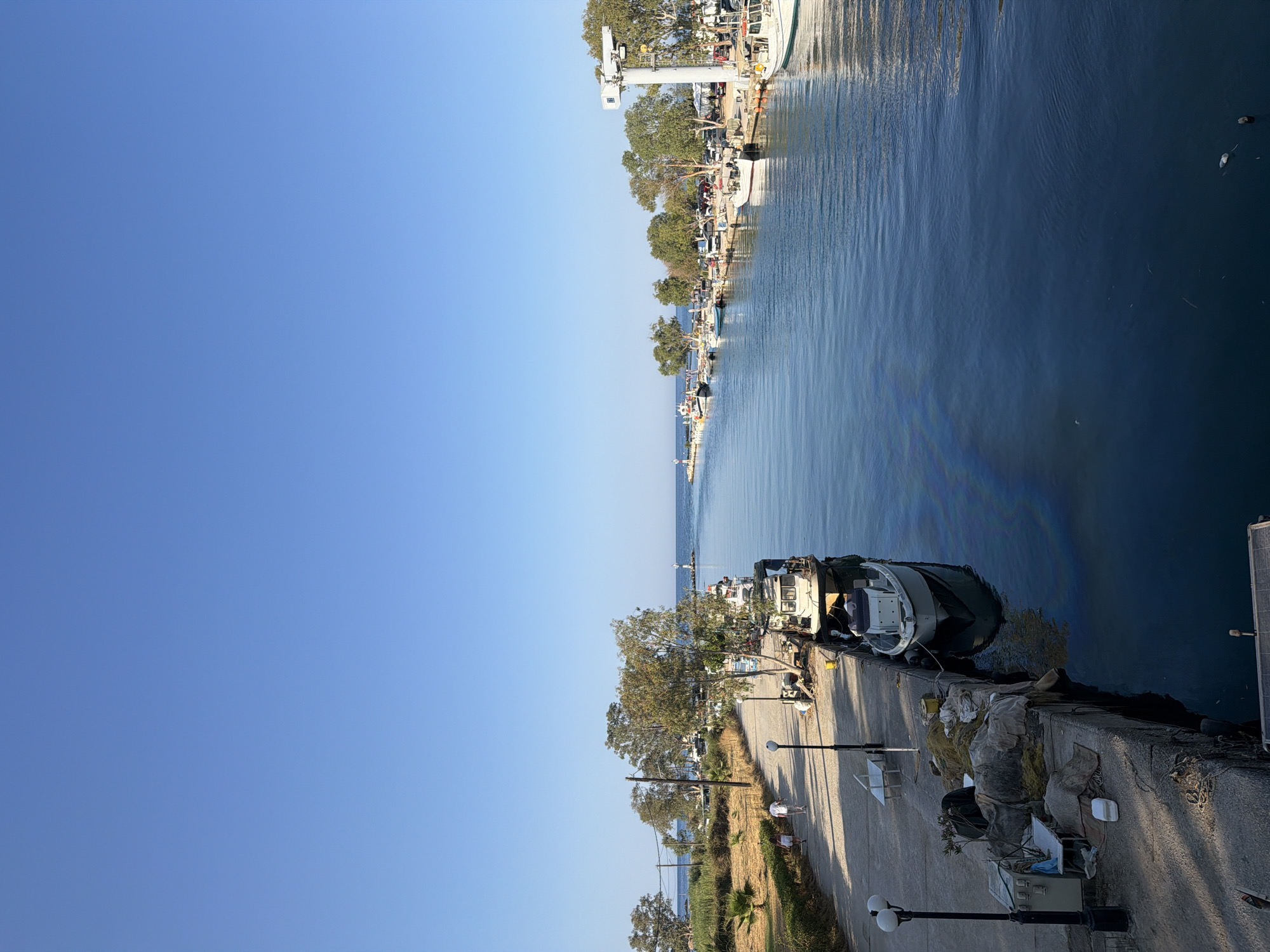 The image shows a view of the harbor in Georgioupoli, Greece, on a clear day with a bright blue sky. The water is calm and dark blue, with subtle rainbow-like iridescence visible on the surface in the foreground. Several boats are docked along a concrete pier on the right side of the image, which extends into the distance. These boats appear to be fishing vessels, with some being smaller and others larger.
On the left side of the harbor, a paved walkway runs parallel to the water. Two people, a man and a woman, are walking away from the camera on this walkway. The area to the left of the walkway is lined with trees, dry grass, and some palm trees. Further down the walkway, a white bench and a lamppost are visible. Fishing nets and equipment are piled on the pier near the lamppost. In the distance, a lighthouse can be seen at the mouth of the harbor, and more boats are docked on the opposite side of the waterway. A large white crane structure is also present in the background, near the edge of the harbor. The overall scene depicts a tranquil coastal town harbor.