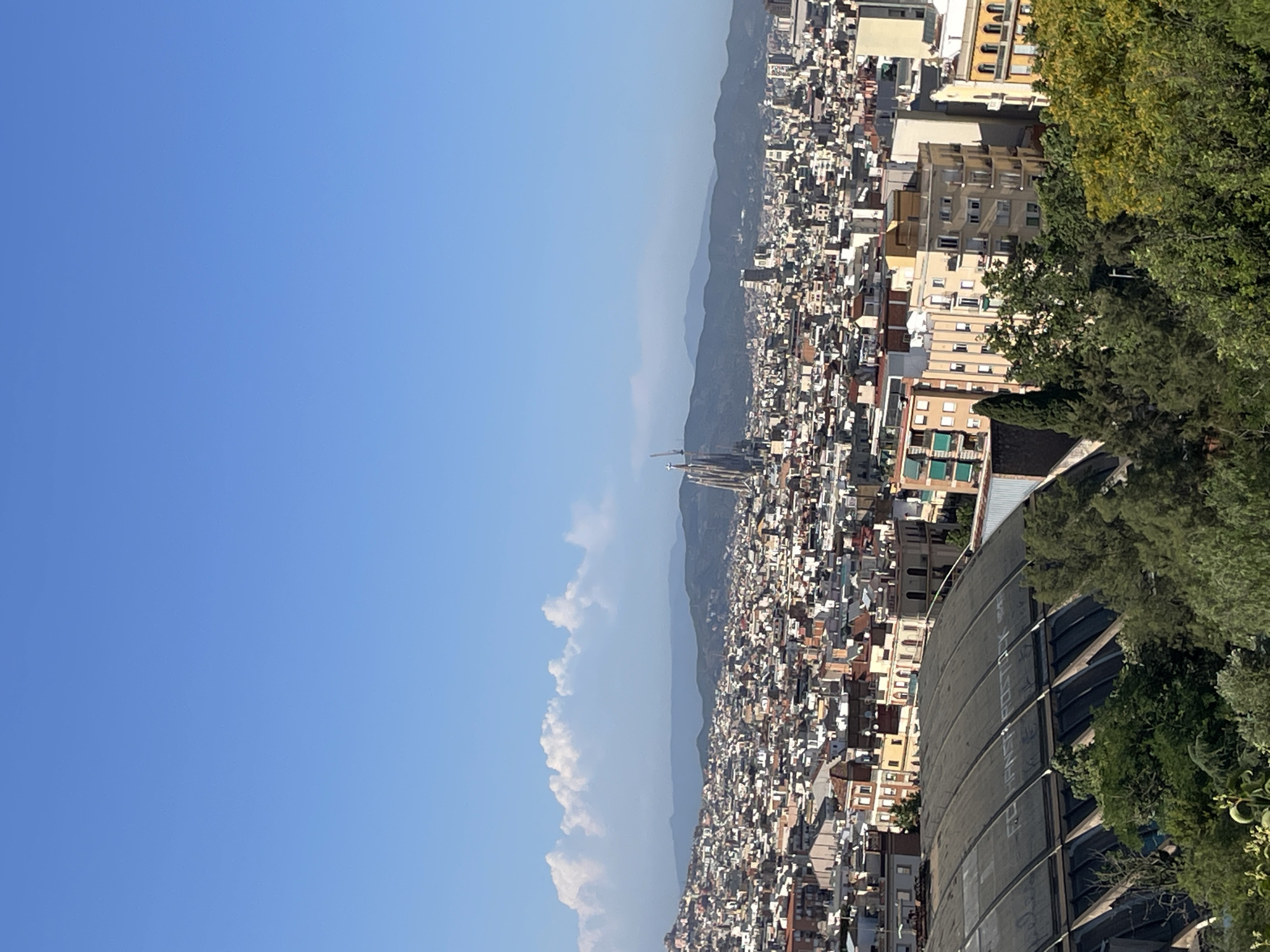 The image is a panoramic view of the city of Barcelona, Spain, on a clear day with a few scattered clouds. The foreground shows a dense canopy of green trees. Behind the trees, a large, arched structure with a dark, ribbed roof is visible, appearing to be a stadium or large event hall. Beyond this structure, the cityscape of Barcelona unfolds, characterized by a dense collection of multi-story buildings with varied architectural styles and rooftop details. In the mid-ground, the iconic Sagrada Familia basilica with its distinctive spires stands prominently amongst the urban sprawl, indicating its location within the city. In the background, rolling hills and mountains form a natural horizon. The sky is a bright, clear blue, with a line of fluffy white clouds stretching across the middle distance. The overall impression is of a sunny day offering a sweeping vista of the city and its surroundings.