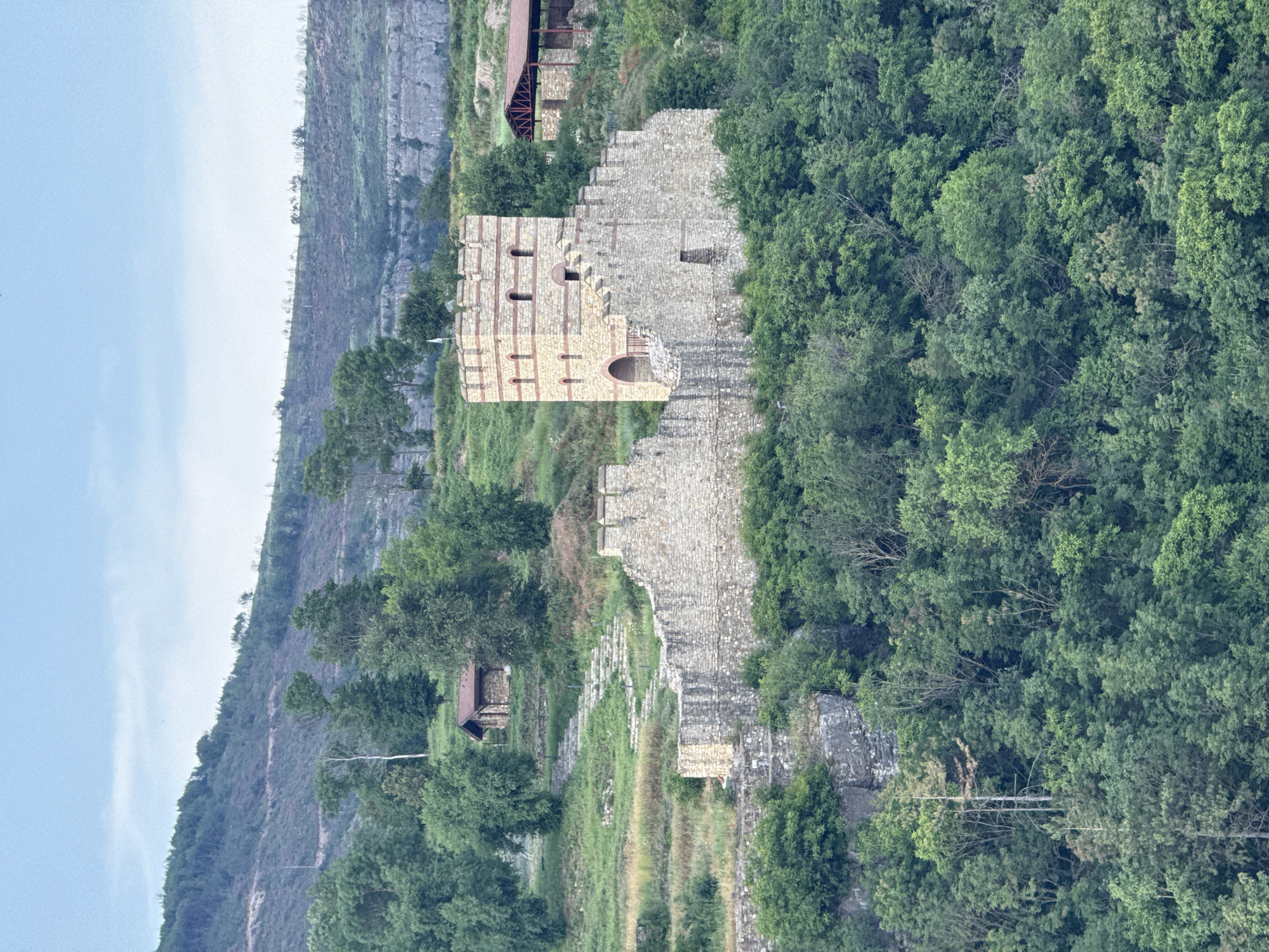 The image depicts a portion of the medieval fortress of Tsarevets in Veliko Tarnovo, Bulgaria, specifically a ruined stone tower and a section of the fortress wall. The tower, made of stone with red brick horizontal bands, has visible openings and some collapsed sections. The wall extends downward along the slope, also in ruins. Lush green vegetation covers the surrounding area, including trees and grass. A rocky cliff face is visible in the background to the left of the tower. The sky is a pale blue.