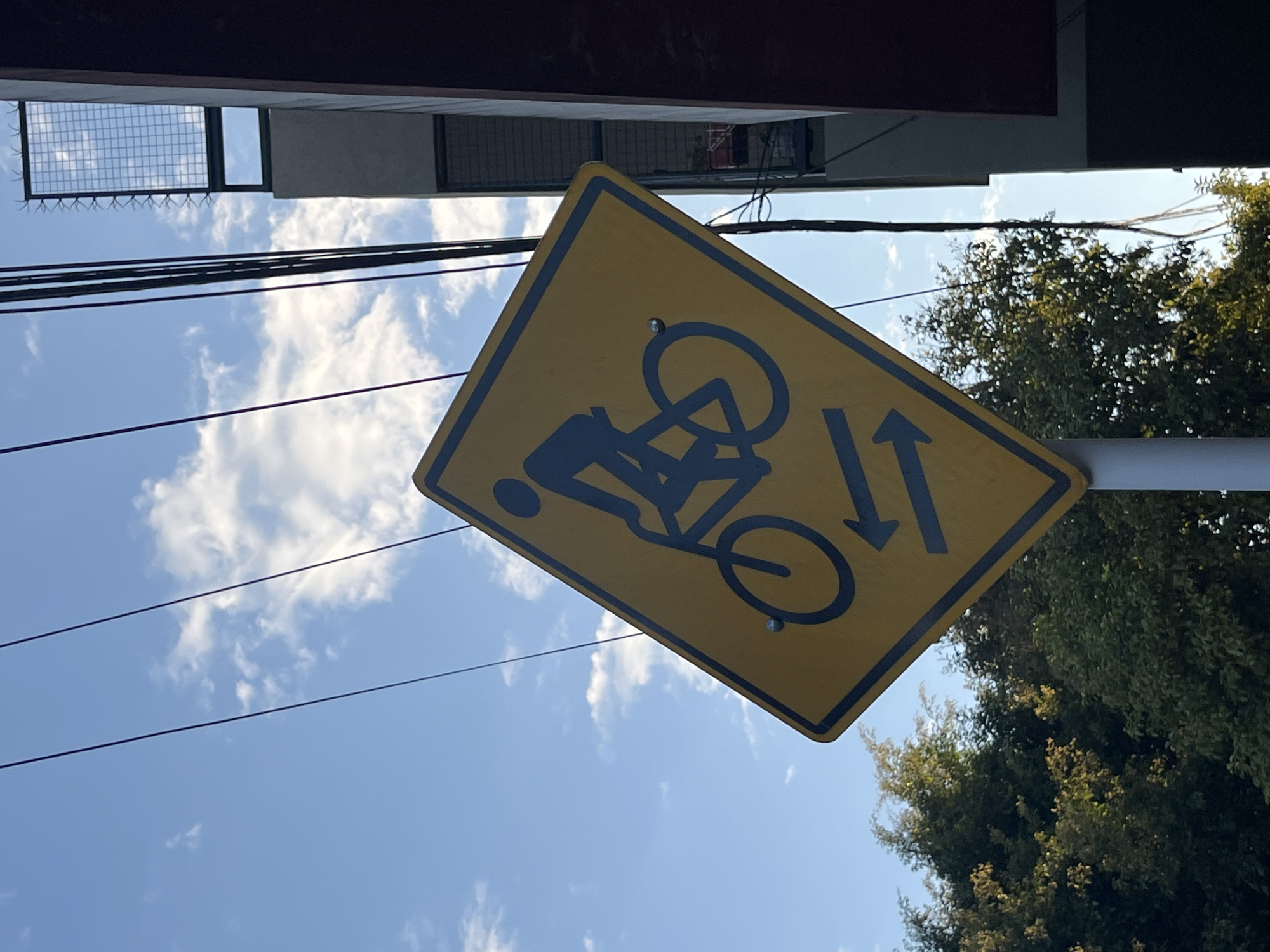 A low-angle shot captures a vibrant street scene looking upwards towards a bright blue sky filled with scattered white clouds. The primary subject is a yellow, diamond-shaped traffic sign, tilted diagonally across the frame, indicating a bicycle path. The sign features blue pictograms: a person riding a bicycle, and two vertical arrows pointing in opposite directions, signifying a two-way bike lane. The sign is affixed to a grey pole visible on the right side.
Above the sign and extending across the sky, several dark electrical wires run horizontally. On the left side of the frame, parts of an urban structure are visible, including a dark building edge and a metal security grid with anti-climb spikes against the sky. On the right, the dense green canopy of trees provides a natural contrast to the urban elements. The top edge of the image shows the underside of an overhanging structure or roof. The clear sky and bright lighting suggest it is daytime. The scene provides a glimpse into a typical urban environment in Buenos Aires, Argentina, emphasizing bicycle infrastructure.