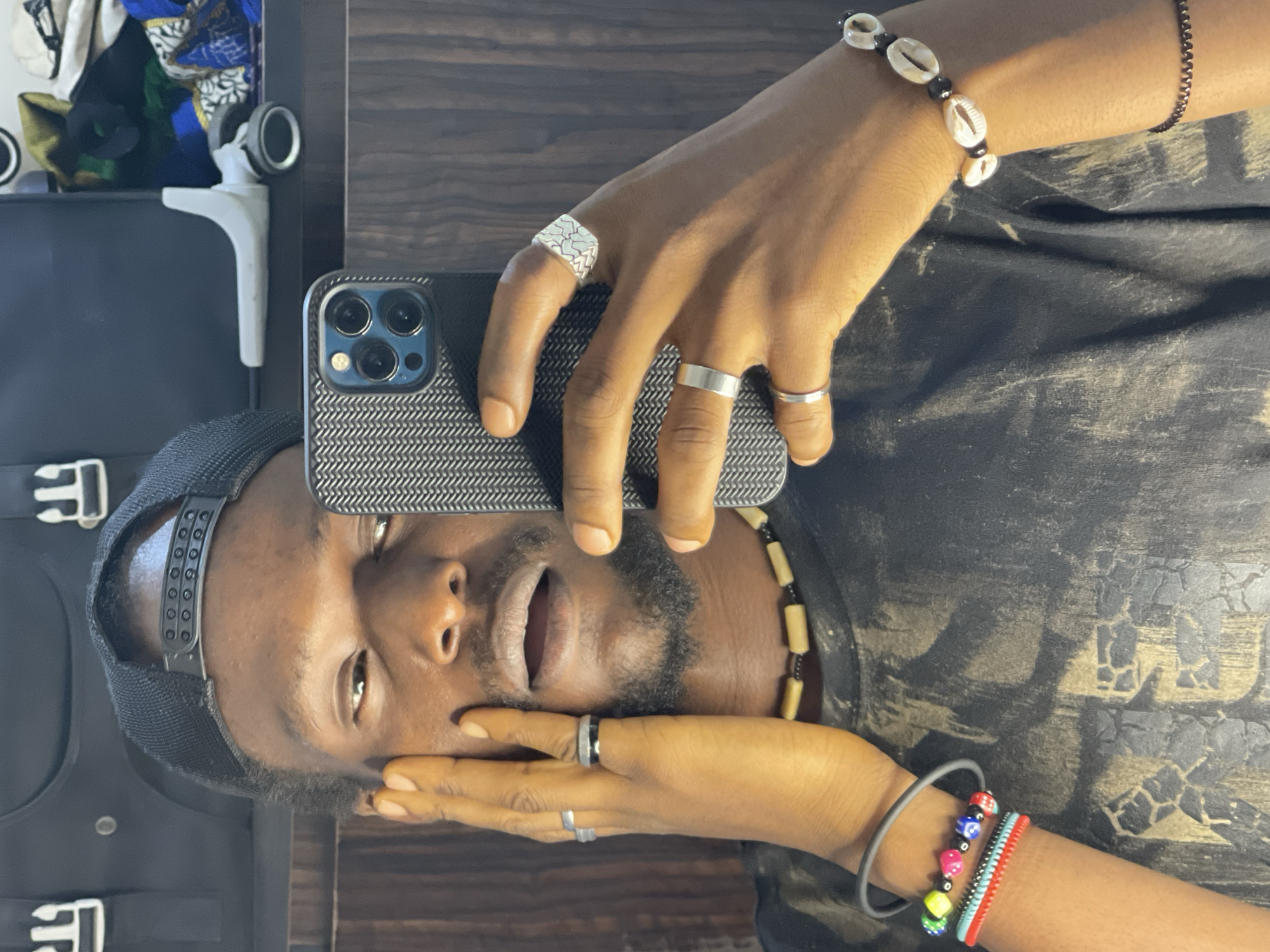 A young Black man is taking a selfie in a dimly lit room with wooden paneling and a dark background that appears to be a closet or wardrobe. He is wearing a black baseball cap and a black t-shirt with a distressed, abstract design in a lighter color. He is also wearing several rings on his fingers and beaded necklaces around his neck and on his wrist. One bracelet features cowrie shells. He is holding a smartphone in his right hand, angled to capture his reflection in a mirror. His left hand is placed on his cheek, and he appears to be looking at the phone's screen with an open mouth. The overall impression is a casual, personal moment. There are no discernible indicators to place the image specifically in Abuja, Nigeria.