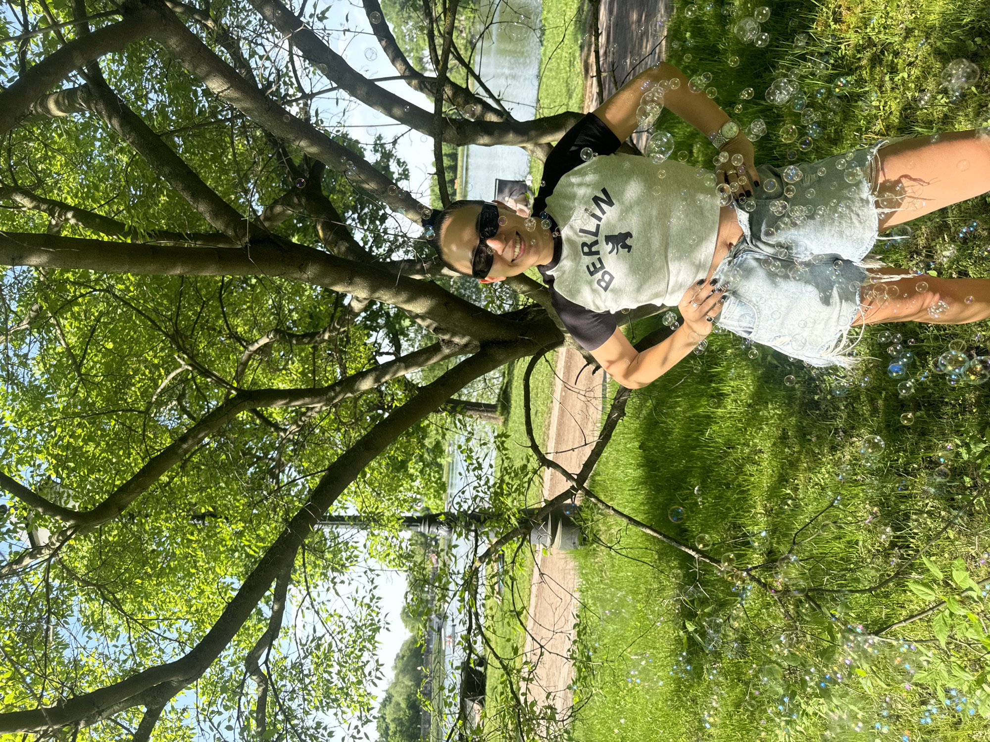 A smiling woman wearing a "Berlin" t-shirt, jean shorts, and sunglasses is posed in front of a tree, surrounded by bubbles, in a park setting. A body of water is visible in the background, and a path runs through the grassy area. The location is in Moscow, Russia.