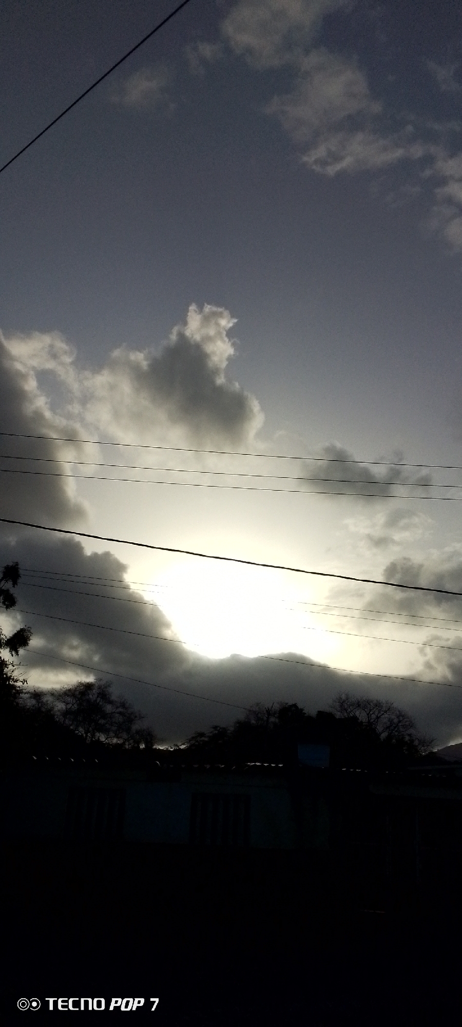 The image, taken in the city of Altagracia, Venezuela, shows a twilight sky with various cloud formations. The sun is partially obscured by clouds, casting a bright glow. Several utility lines stretch across the frame, partially obscuring the sky. Below the horizon line, silhouetted trees and what appear to be buildings are visible. The scene is mostly dark, with limited details visible in the foreground due to low light. The bottom of the frame contains the phone model information "Tecno Pop 7".