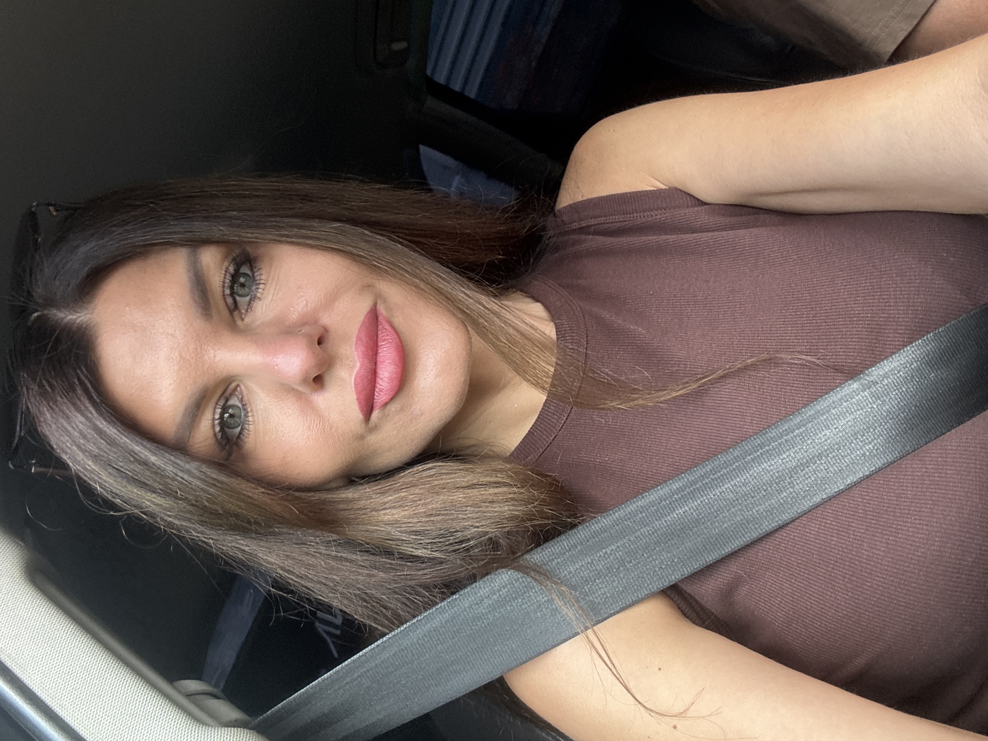The image is a close-up selfie of a woman sitting in a car, with a seatbelt prominently displayed across her chest. She has long, brown hair and is wearing a brown, sleeveless, ribbed top. The background is dark and out of focus, suggesting the interior of a vehicle. The woman's face is centered in the frame, with her gaze directed towards the camera. She is wearing makeup, including lipstick. The environment appears to be within a car. There is no indication of the specific location within the city of Chaikovsky, Russia.