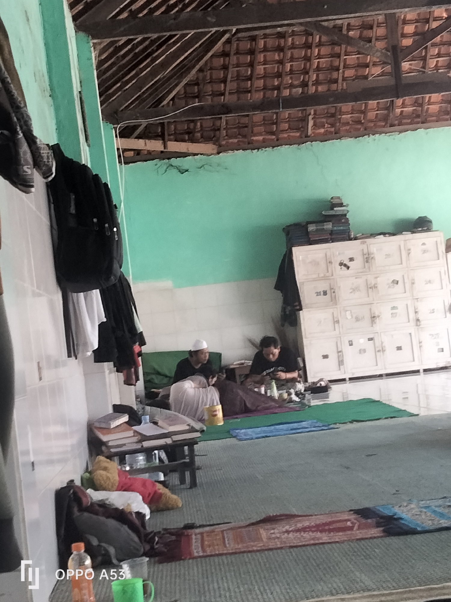 The image depicts an interior scene, likely within a building in Badung, Indonesia. The room has a high, pitched ceiling with exposed wooden rafters and terracotta tiles. The walls are painted a light green. In the foreground, a light-colored tiled wall is visible on the left.
On the right side of the image, a large, white, multi-compartment cabinet or locker unit is present, with several numbered drawers or doors, suggesting it could be for storage or personal effects. Books are stacked on top of this cabinet. In front of the cabinet, two individuals are seated on the floor. One person, wearing a white skullcap, is looking to the left. Another person, wearing a black t-shirt, is looking down and appears to be engaged with something on the floor. Several items, including bottles and containers, are scattered around them.
In the middle ground, a low, dark table holds a stack of books. To the left of the table, a green mat is laid out, with a plush toy on it, wearing a red garment. Several bags are also present, including a black backpack hanging on the left wall. On the floor, prayer mats are visible, one a muted red and blue pattern, and another green mat.
In the bottom left corner, a plastic bottle of orange liquid and a green plastic mug are visible. The logo "OPPO A53" is also present in this corner, indicating the device used to capture the image. The overall scene suggests a communal space, possibly a place of worship or a dormitory, given the presence of prayer mats and storage units.