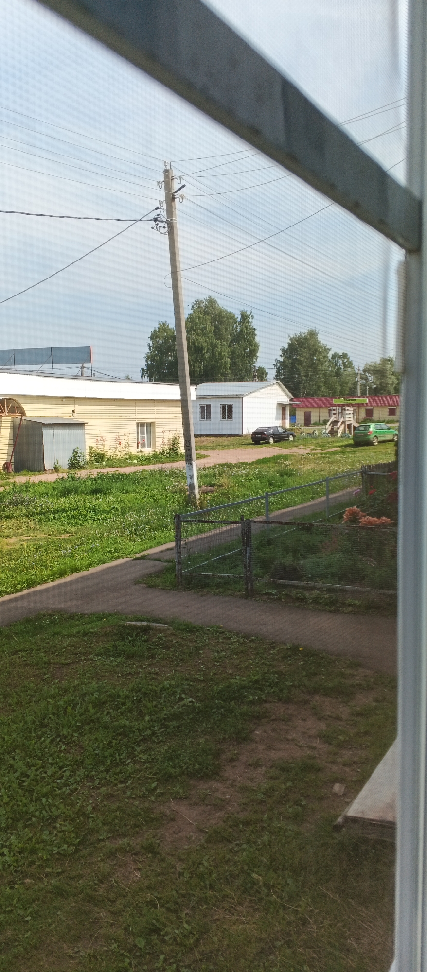 The image shows a view through a window screen of a rural or suburban area in the village of Podgornoye, Russia. In the foreground, there is a grassy area with a paved walkway. A metal fence runs along the walkway. In the middle ground, there are several low-rise buildings. One building is yellow with a white roof, and another is white with a red roof. There are trees in the background. Two cars are parked in front of the buildings: a dark-colored sedan and a green hatchback. A utility pole with wires is visible to the left of the frame. The sky is light blue with some clouds.