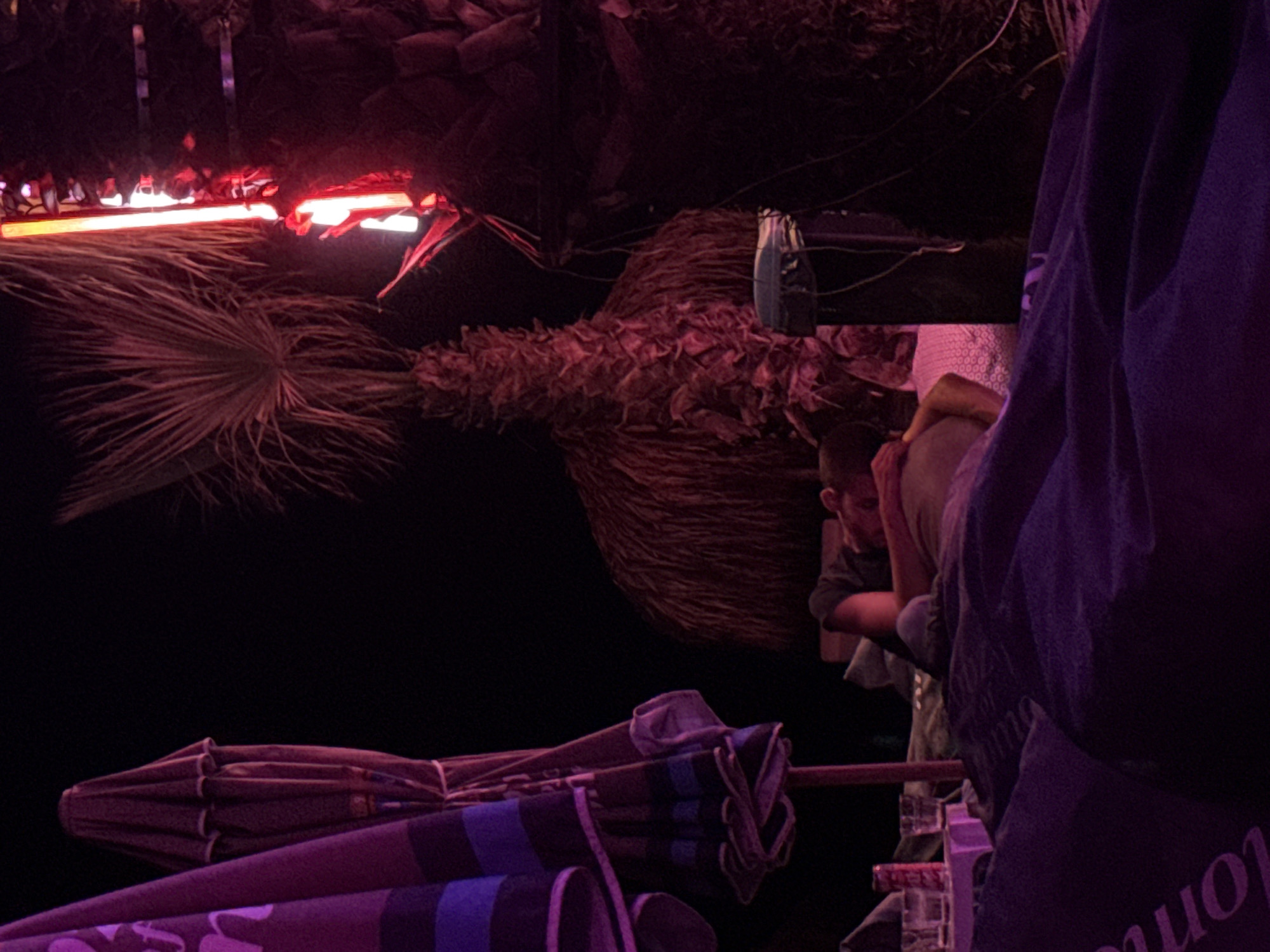 The image depicts a nighttime scene at what appears to be an outdoor venue with a beach or tropical theme, possibly in Burgas, Bulgaria. In the foreground, a person is lying down, partially covered by a dark purple blanket or fabric with indistinct text. Their back is to the camera, and they are resting on pillows or cushions. Further back, a person is seen from the side, also lying down, with their head resting on their arm. They are wearing a dark shirt.
To the left of the frame, two beach umbrellas are visible, one purple with blue stripes and the other a darker color. They are closed and appear to be made of fabric. Behind the umbrellas, the dark night sky is visible.
In the background, a thatched roof structure, possibly a hut or a bar, is present. There are palm trees with distinctive fronds and trunks, illuminated by red and orange neon lights. One neon light strip is clearly visible, casting a bright glow. A trash can or bin with a lid is also visible near the thatched structure. On a small table or surface, there are several glasses and a red soda can. The overall lighting is dim, with strong colored illumination from the neon lights. The environment suggests a relaxed, possibly party atmosphere.