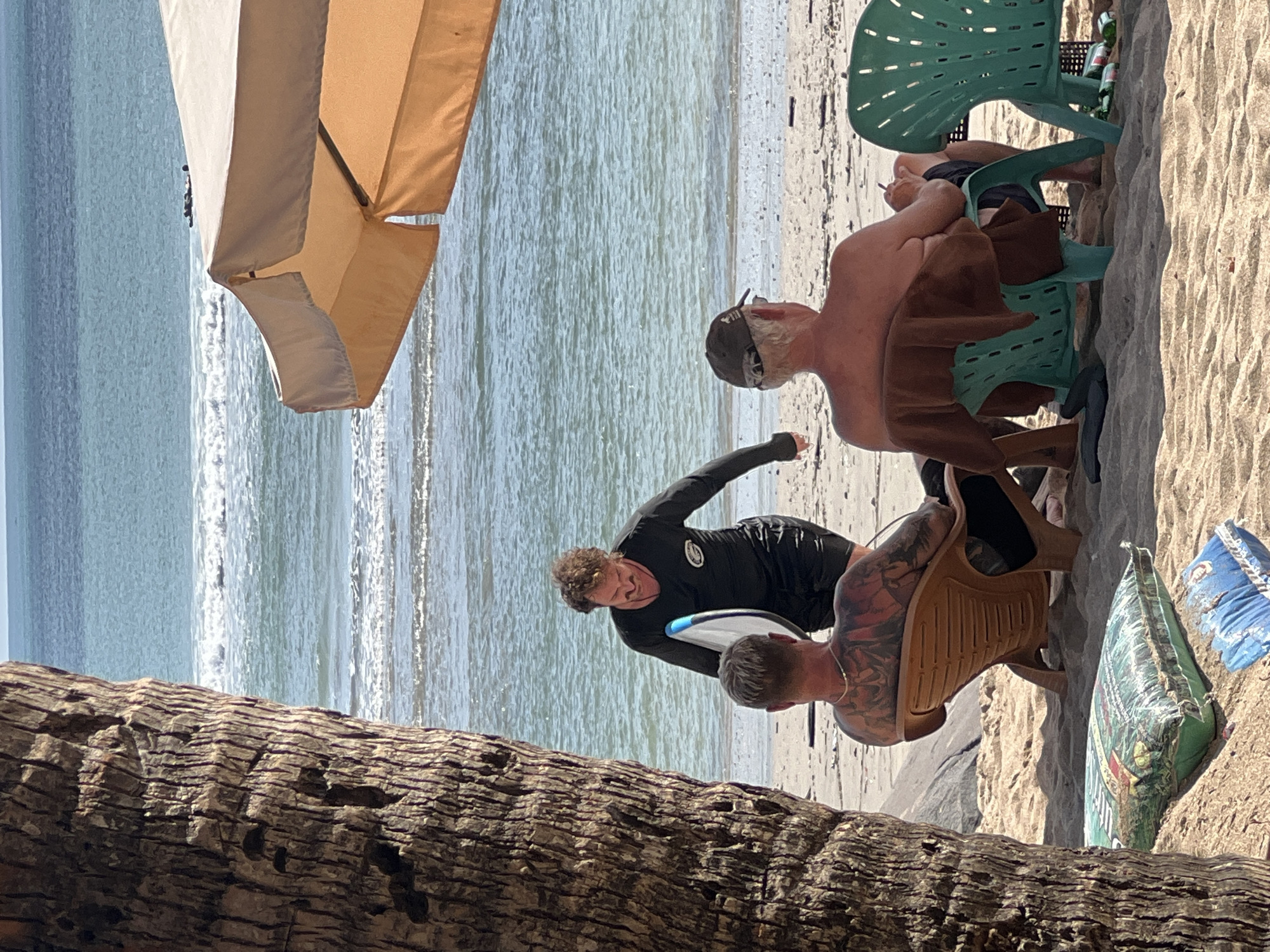 The image depicts a beach scene in Kuta, Indonesia. In the foreground, a large palm tree trunk occupies the left side of the frame. Two men are seated in beach chairs on the sand, facing away from the camera towards the ocean. The man on the left has extensive tattoos on his back. The man on the right is wearing a baseball cap and has a brown towel draped over the back of his chair. Between them, a third man wearing a black long-sleeved rash guard and black shorts is walking towards the seated men, carrying a surfboard. He is smiling and appears to be interacting with them. In the background, the ocean stretches to the horizon, with small waves breaking on the shore. An orange beach umbrella is partially visible in the upper right corner of the image. Several plastic bottles and a bag of what appears to be soil or fertilizer are on the sand.