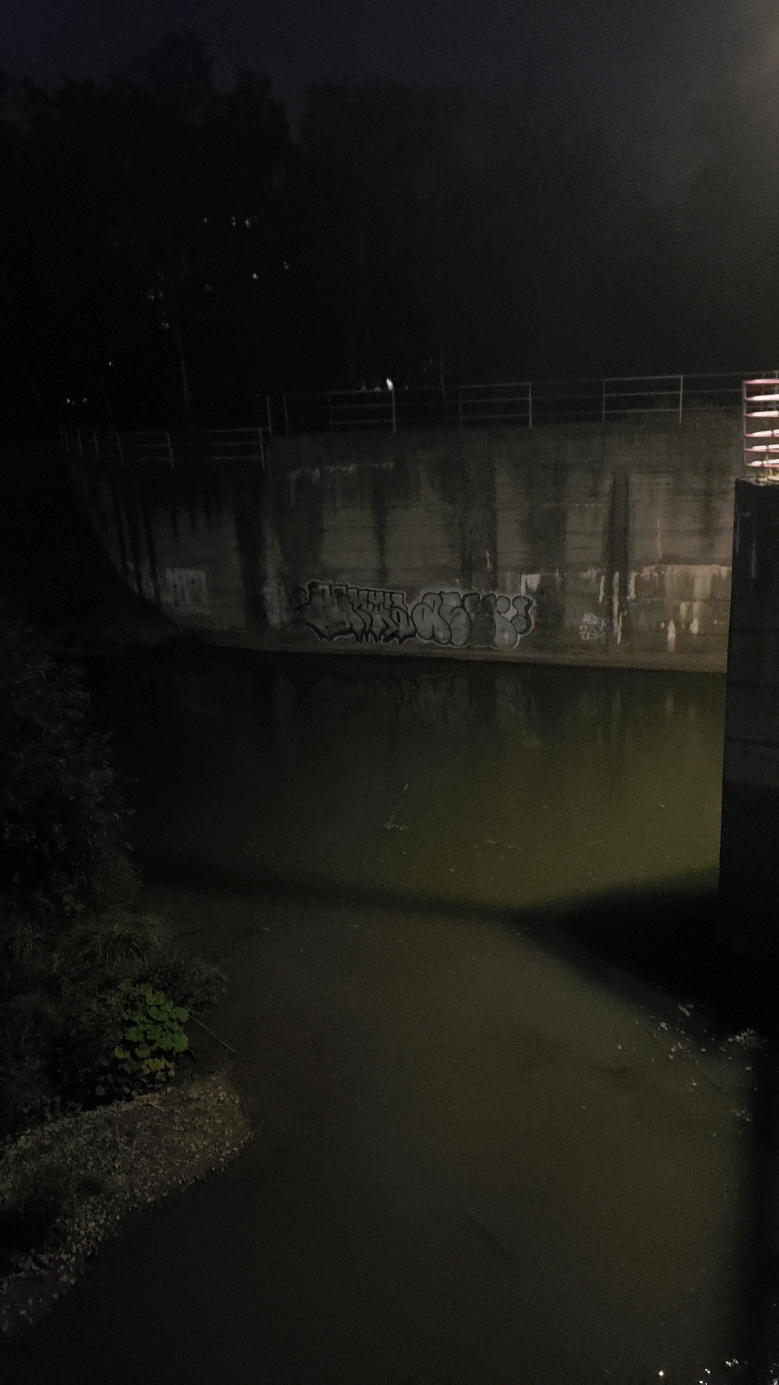 The image depicts a nighttime scene along the Torysa river in Prešov, Slovakia. A concrete embankment lines the river, with graffiti visible on its surface. A metal railing runs along the top of the embankment. Trees are visible in the background, silhouetted against the dark sky. The water of the river is dark and appears murky. Vegetation grows along the left bank of the river. The scene is dimly lit, suggesting the presence of artificial lighting or moonlight.