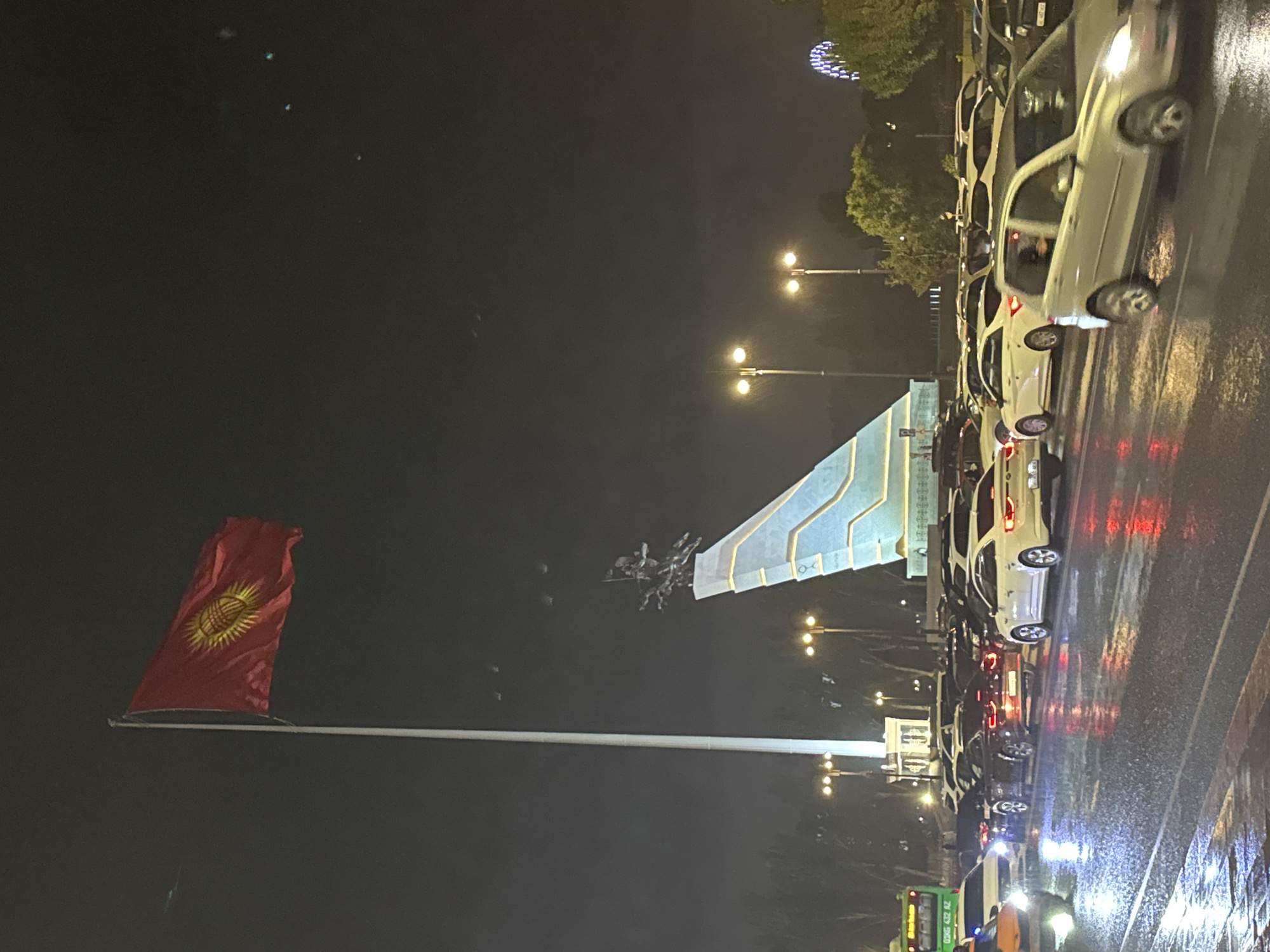 A Kyrgyzstan flag is prominently displayed on a tall flagpole at Ala-Too Square in Bishkek City, Kyrgyzstan, during nighttime. The flag is red with a yellow sun emblem in the center. In the background, a monument featuring a sculpture of a rider on a horse stands atop a modern, angled structure. The square is wet from rain, reflecting the lights of streetlamps and vehicles. A traffic jam is visible, with numerous cars parked and moving on the wet asphalt. Trees line the sides of the square, and a Ferris wheel can be seen in the distance. A green bus with license plate "036G 432 AZ" is also present in the scene. The overall atmosphere is somber due to the nighttime setting and rainy weather.