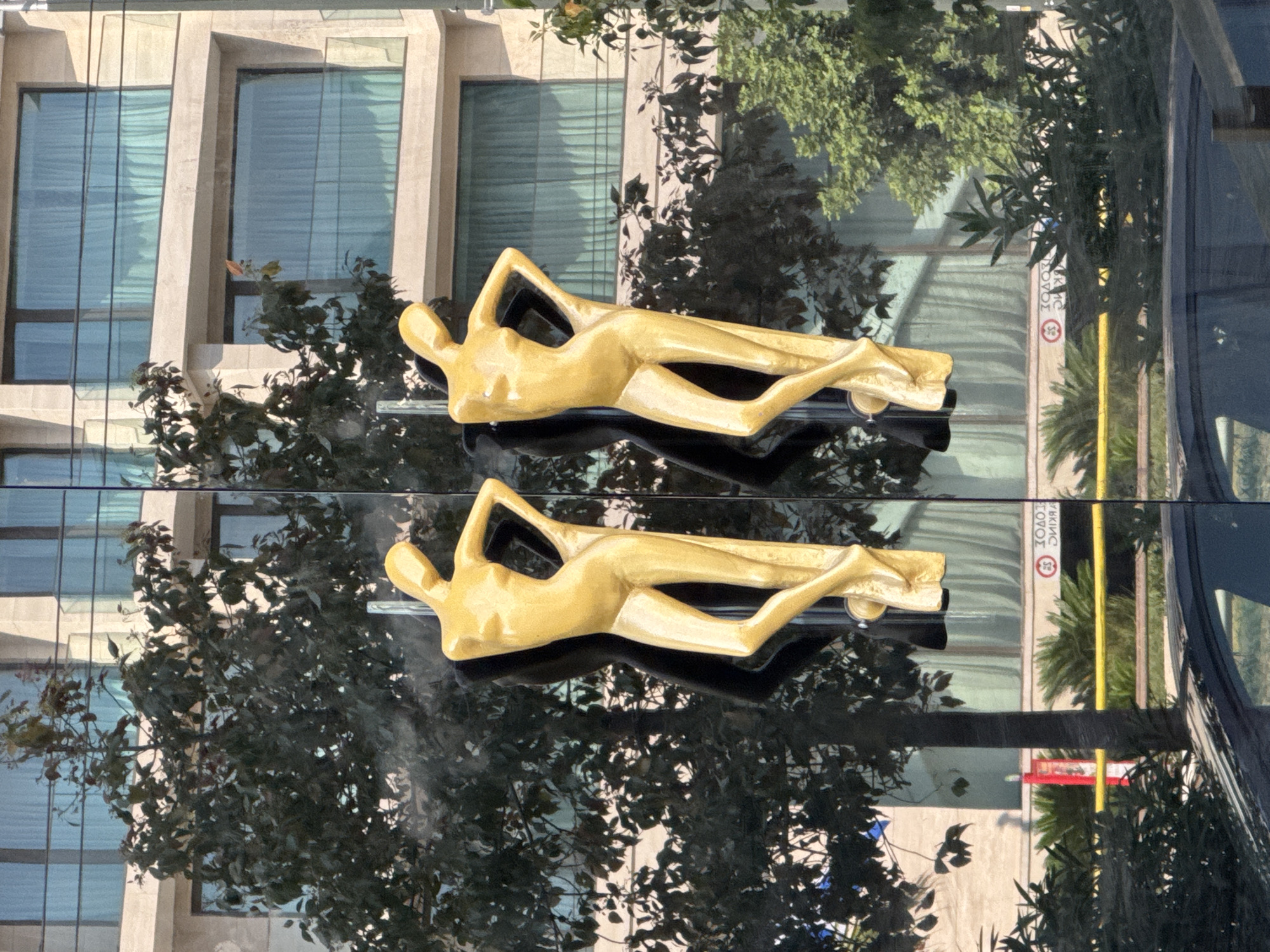 The image shows two gold humanoid sculptures on a set of glass doors. The sculptures are stylized and appear to be female figures. They are attached to the glass doors, and one on the left has a black silhouette behind it. Behind the doors, a modern building with multiple floors and large windows is visible. Trees with dark green foliage are in front of the building and partially obscuring it. In the reflection on the glass doors, there is a car and some greenery. There are also some circular signs with text and symbols visible in the reflection, possibly indicating parking restrictions. The overall scene suggests an outdoor setting in a city, likely a commercial or public space. The presence of modern architecture and the style of the sculptures point to a contemporary urban environment in Athens, Greece.