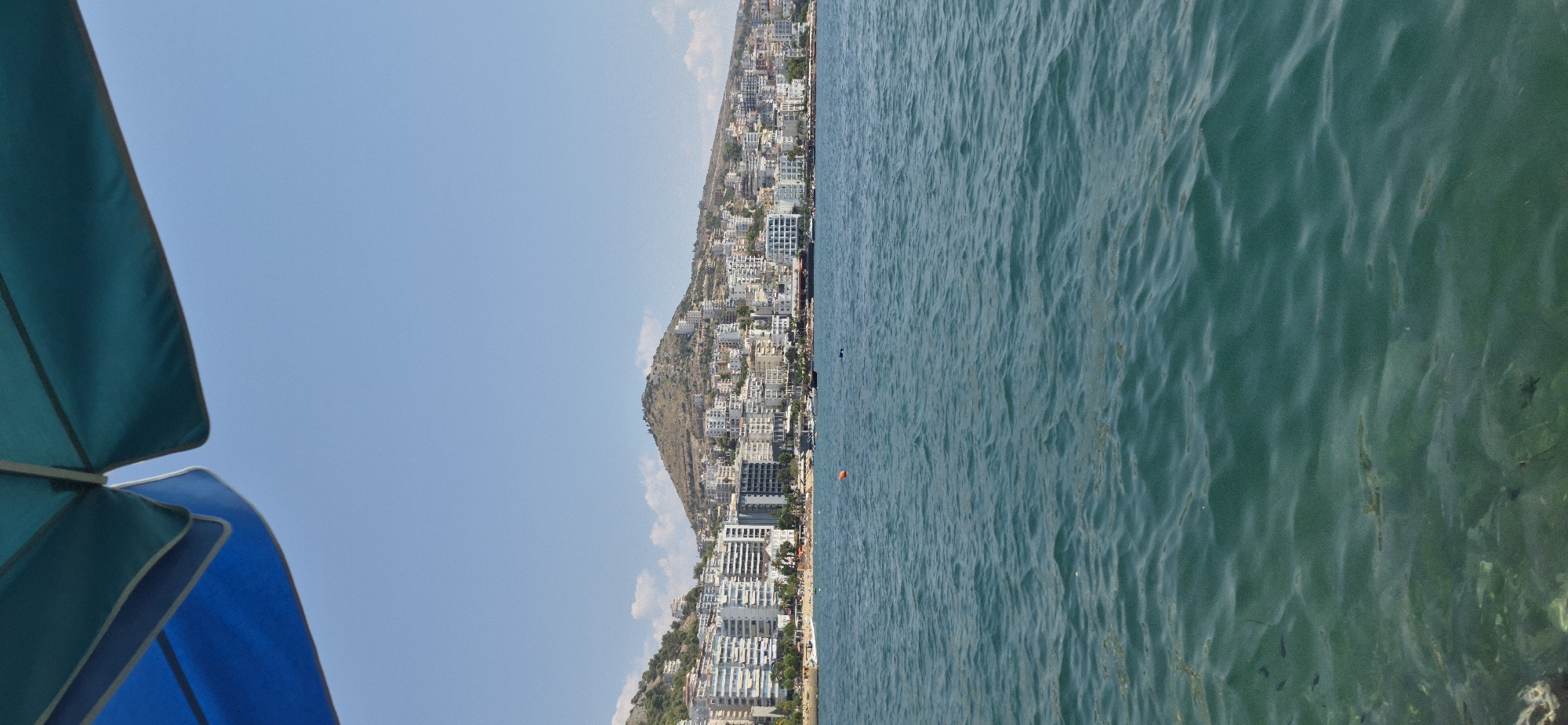 The image shows a view of the city of Saranda, Albania, from the water. In the foreground, the turquoise water is clear, revealing the seabed with rocks and vegetation. Small fish are visible swimming in the water. In the mid-ground, a line of white buildings stretches along the coastline. These buildings are of various heights and styles, with some appearing to be apartment complexes and hotels. A beach area is visible between the buildings and the water, with a few scattered beach umbrellas and sunbathers. Further back, a large hill dominates the landscape, covered in smaller buildings and vegetation. The sky is a clear blue with a few scattered white clouds. The top of the image is partially obscured by a blue and teal umbrella.