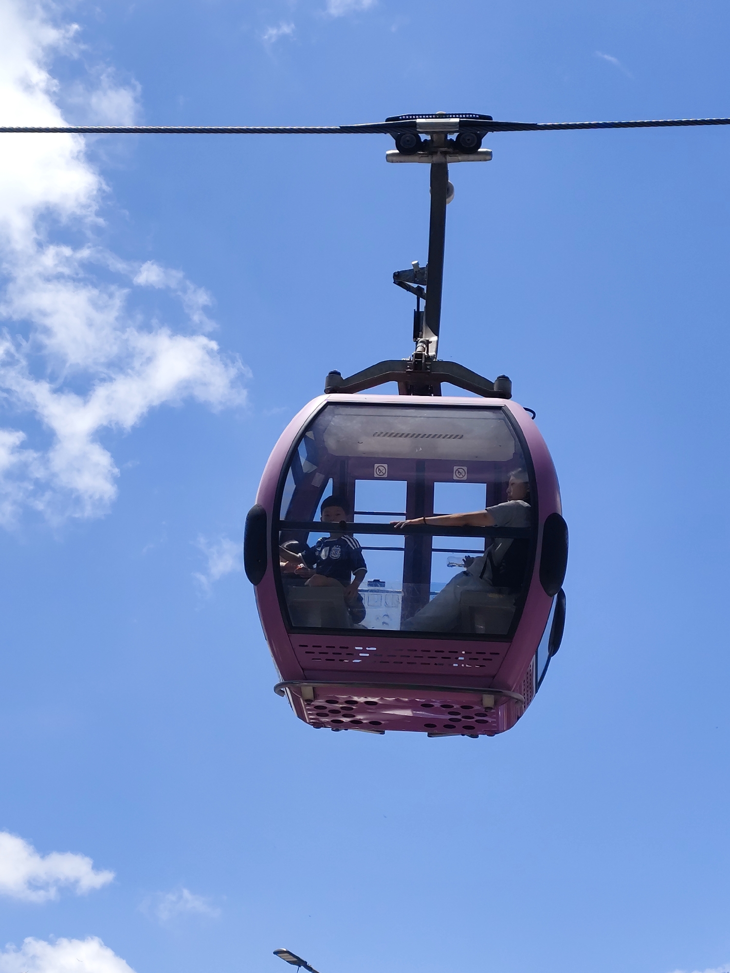 A pink cable car is suspended against a clear blue sky with scattered white clouds. Inside the cable car, two individuals are visible. A young boy wearing a dark blue t-shirt with white accents is seated on the left side. To his right, a woman with light-colored clothing is seated, her left arm extended forward, holding a white object. The cable car is supported by a metal structure attached to a thick cable overhead. The scene is likely in Da Lat, Vietnam, which is known for its cable car systems that offer scenic views of the surrounding landscape.