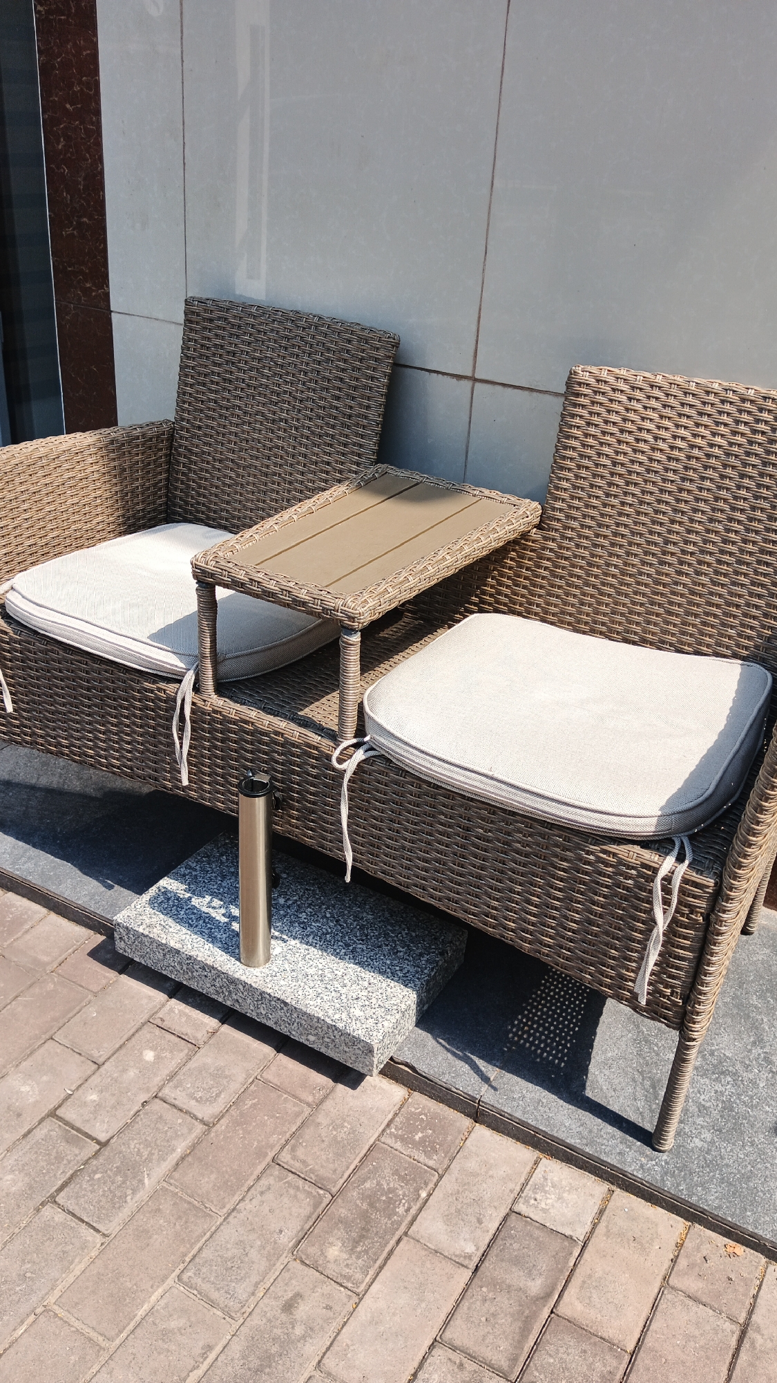 This is a medium shot, outdoor photograph of a wicker loveseat with a small table between the seats. The loveseat has two light-colored cushions. A silver water bottle stands on a gray granite block to the left of the loveseat. The loveseat is on a gray paved patio, which is in front of a building with a tiled facade. The scene is set in the city of Kryvyi Rih, Ukraine.