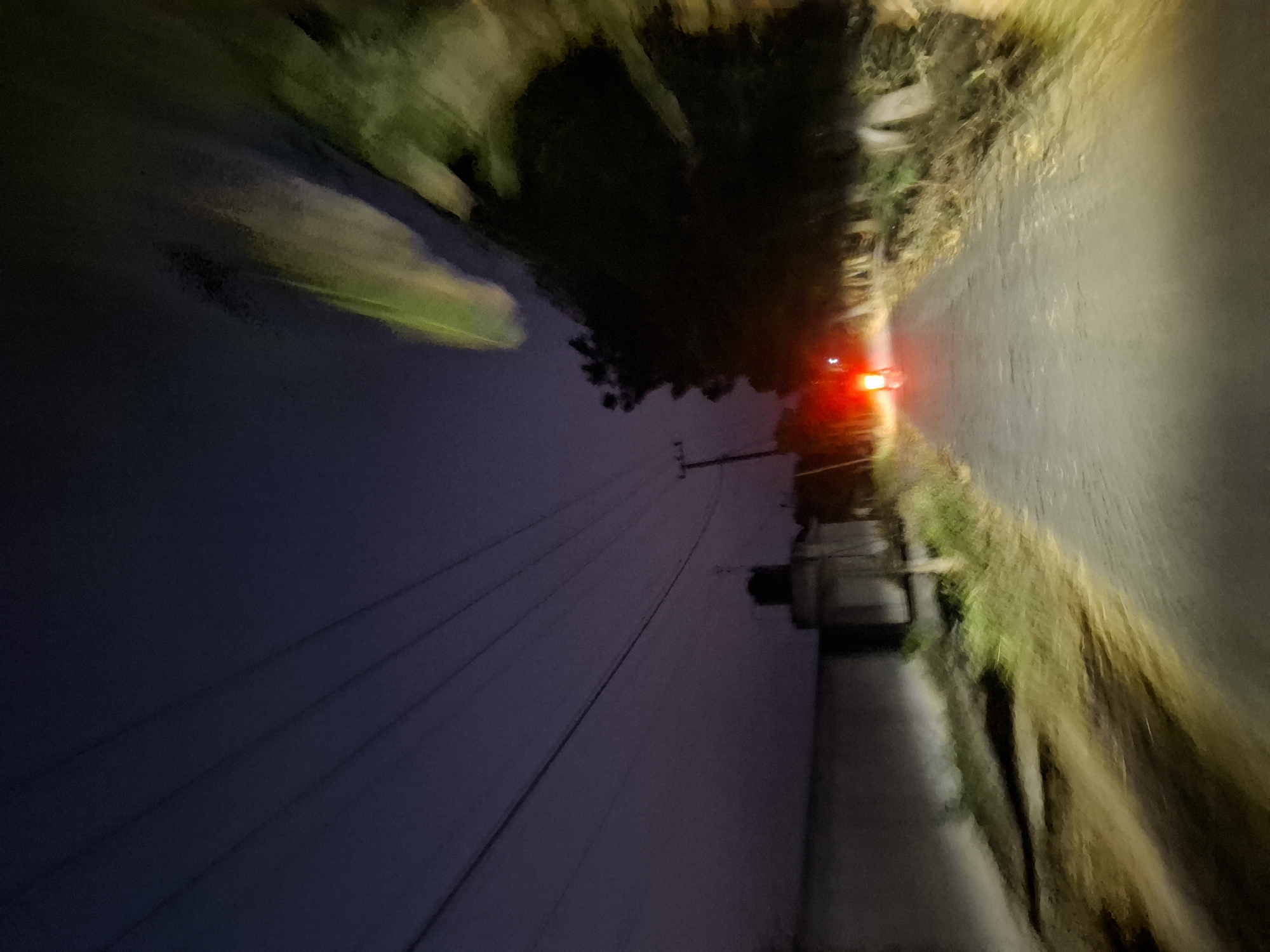 The image depicts a road at night in Bulubrangsi, Indonesia, viewed from the perspective of a moving vehicle. The road is paved and appears to be a two-lane street. In the distance, the red taillight of a motorcycle is visible, indicating another vehicle is ahead. Trees line the right side of the road, and foliage and dry grass are present on the verges. On the left side, a low wall or structure is visible, adjacent to a darker, textured surface. Overhead, power lines stretch across the dark, twilight sky. The overall impression is one of a rural or semi-rural road at dusk or night.