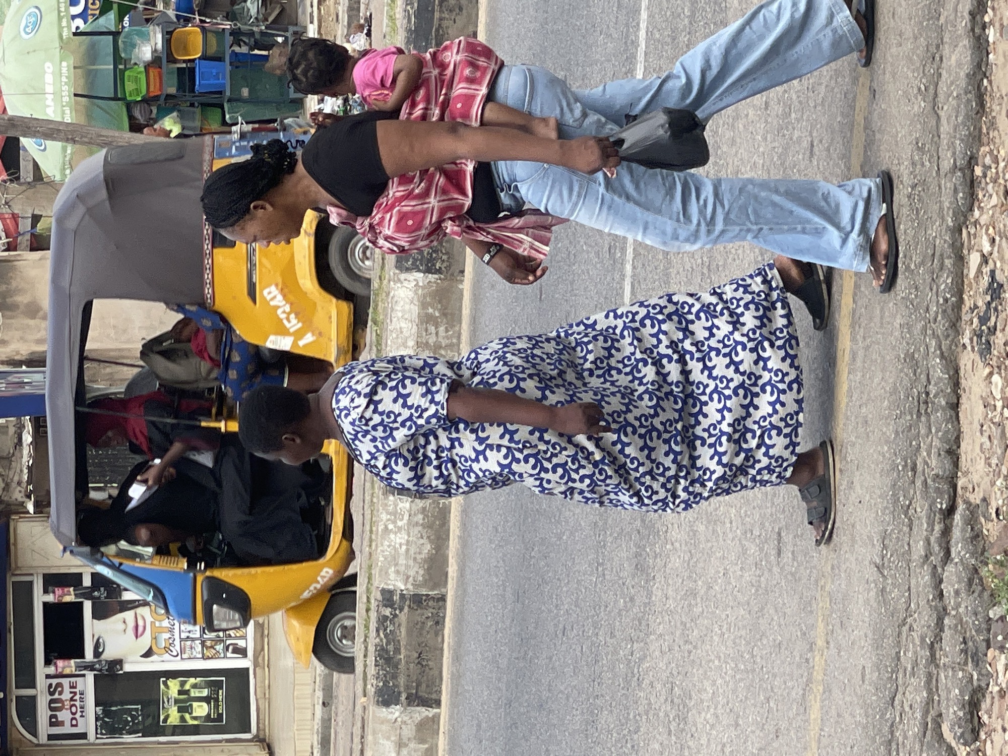 This vertical, eye-level, medium shot captures an outdoor scene on a paved road, likely in a tropical or sub-tropical environment, indicated by the presence of a brightly colored yellow auto-rickshaw and the general street activity. The sky is not visible, so the time of day and weather are indeterminate.
The primary subjects are three individuals, two adults and a child, positioned on the right side of the frame, walking away from the viewer. The person in the foreground, closest to the camera, is wearing a blue and white patterned dress and dark sandals. To their right and slightly behind is another adult, who appears to be carrying a child. This person is wearing a black t-shirt, light blue jeans, and dark sandals, with a red and white checkered cloth wrapped around their torso, securing the child who is also wrapped in a similar cloth. The child is wearing a pink shirt.
A prominent yellow auto-rickshaw, a common form of public transportation in many parts of the world, is parked on the left side of the road. The auto-rickshaw has a grey canopy. The side of the auto-rickshaw features some text and graphics, including what appears to be a large image of a woman's face with makeup, along with text that reads "Cosmet." Another visible sign on the front of the auto-rickshaw displays text including "POS IS DONE HERE" and "SOLD HERE." To the left of the auto-rickshaw, there is a stall with various items, including brightly colored plastic containers and what looks like a green umbrella with "AMEBO Dial *555*PING" and "The No. 140" printed on it.
The scene suggests a bustling urban or semi-urban environment with commercial activity. The focus is on the people and their mode of transport, highlighting everyday life in this location.