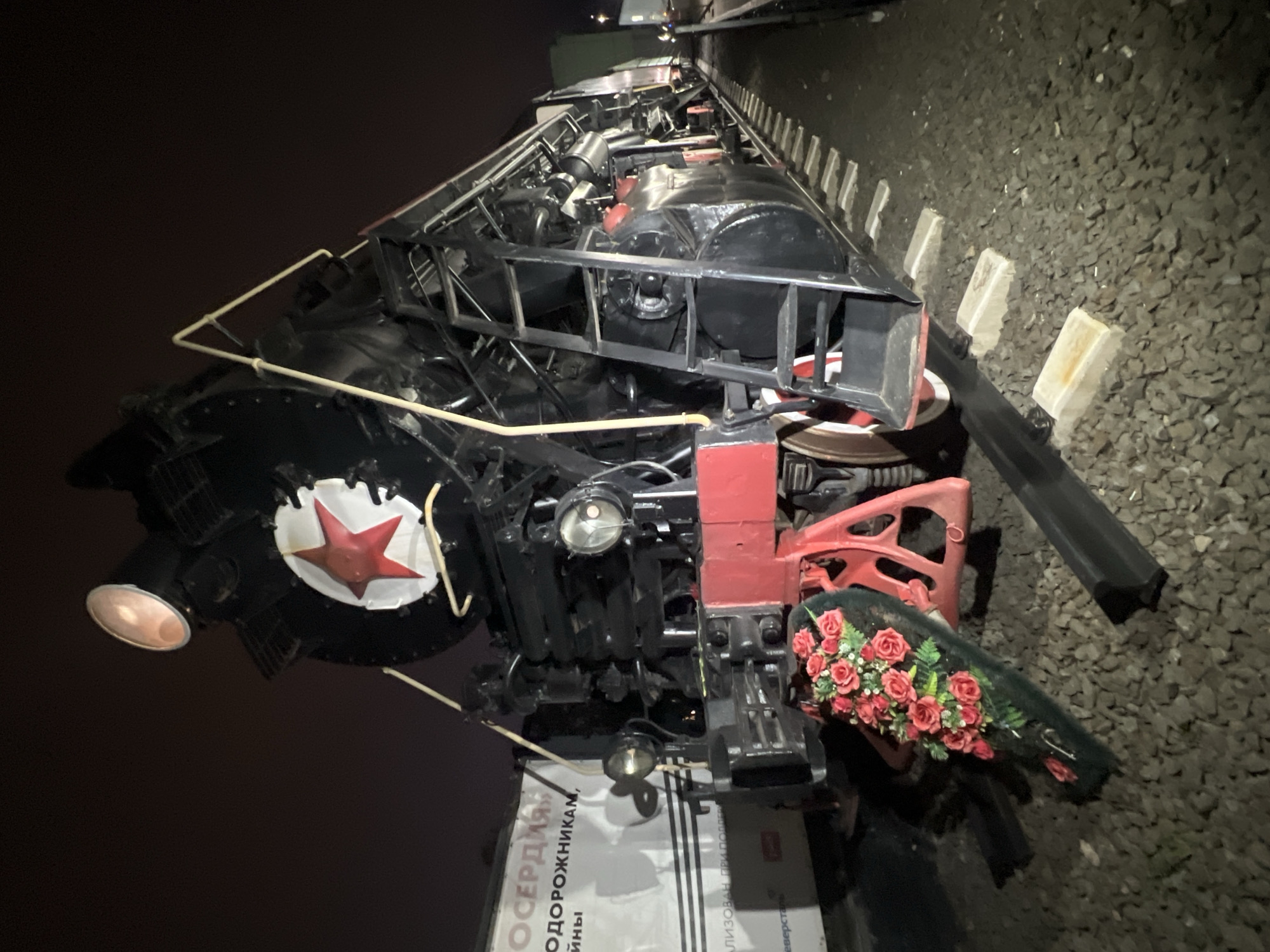 The image shows a detailed view of a black and red Soviet-era steam locomotive, complete with a red star emblem, positioned on railroad tracks. The photograph seems to be taken at night, given the dark background. The train is adorned with a floral arrangement of red roses, seemingly placed as a memorial. A sign with Russian text is visible near the base of the train. The locomotive is located in the city of Cherepovets, Russia. The image is shot from an unusual angle which emphasizes the height of the locomotive.