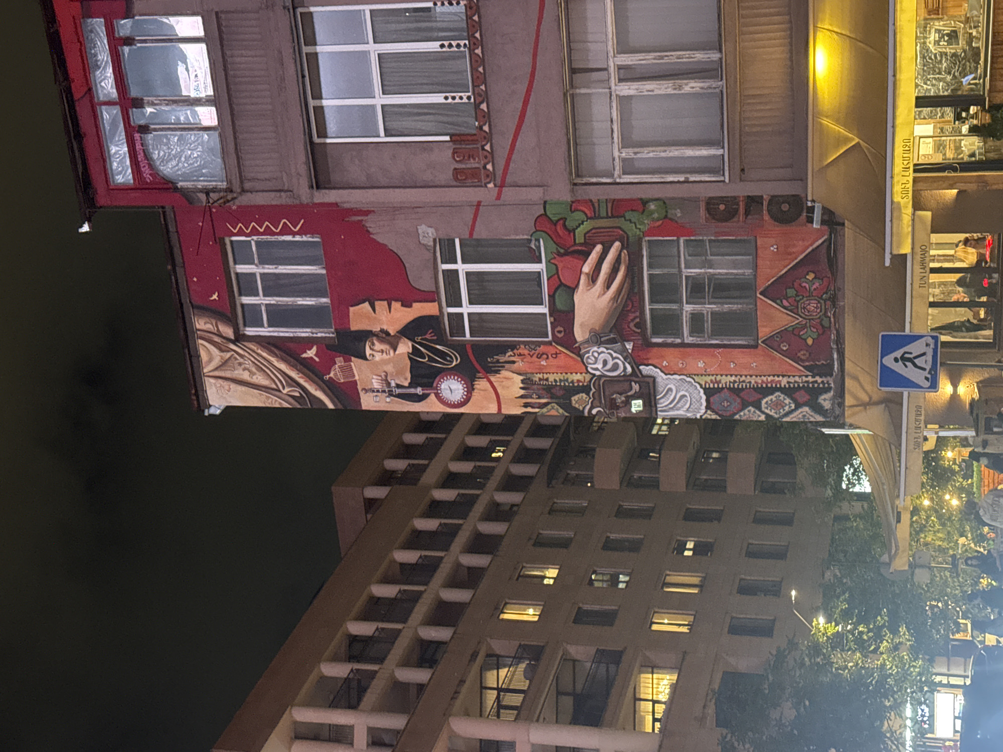 The image depicts a street scene in Yerevan, Armenia at night. A building with a large, colorful mural is prominently featured. The mural depicts various figures and patterns, including a man, a hand, and carpet designs. Adjacent to this building is another high-rise building with numerous windows, some illuminated. A pedestrian crossing sign is visible, and a building with an awning, lights, and signage in Armenian script suggests the presence of a commercial establishment. The environment appears urban, with buildings of varying heights and architectural styles.