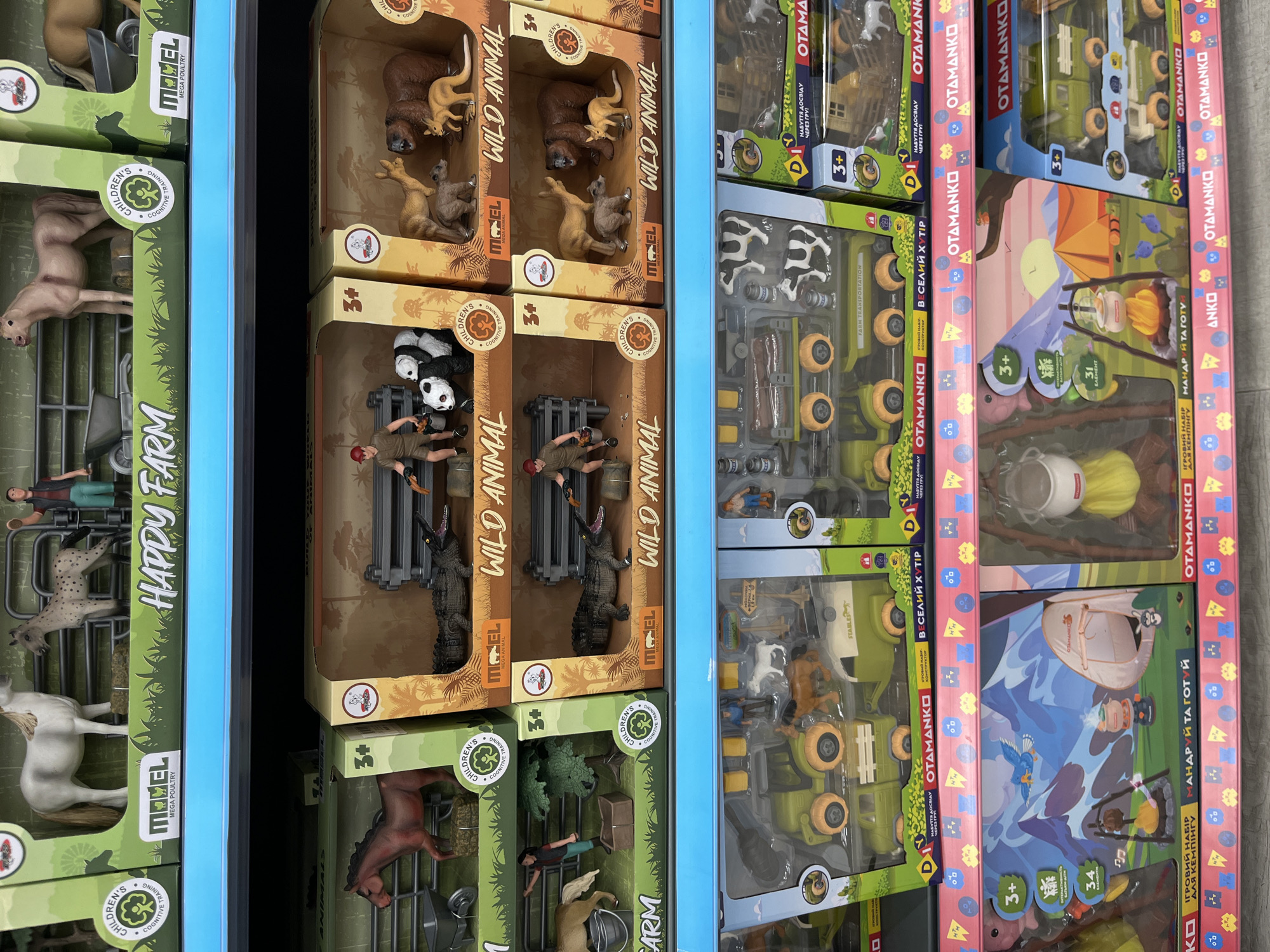 The image shows a retail display shelf in a store, likely a toy store or a department store, in Kharkiv, Ukraine. The shelf is stocked with various toy farm animal and camping sets. The toys are packaged in colorful cardboard boxes, with some boxes featuring illustrations of animals like horses, crocodiles, pandas, bears, and kangaroos. Other boxes showcase farm vehicles like tractors and trailers, as well as camping scenes with tents and campfires. There are no people visible in the image. The lighting appears to be artificial, typical of an indoor retail environment. The overall impression is of a well-organized display of children's toys.