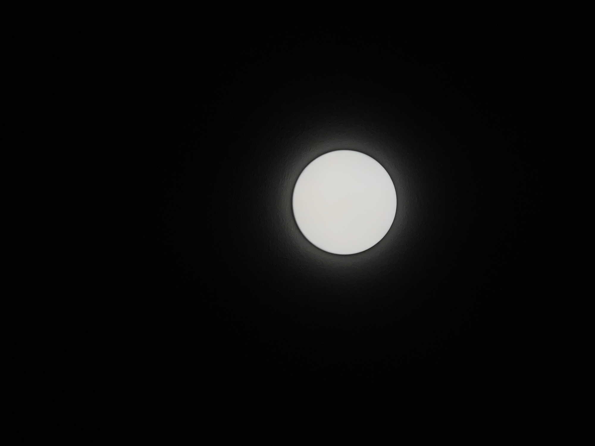 The image displays a circular, white light fixture against a dark, textured background. The light is centered vertically and horizontally in the frame, emitting a soft glow that creates a subtle halo effect on the surrounding surface. The background appears to be a wall or ceiling, with a visible texture that is most pronounced around the light. No people, animals, or other objects are present in the frame. The scene is devoid of any specific indicators that would place it in Tbilisi, Georgia; it is a generic interior shot.