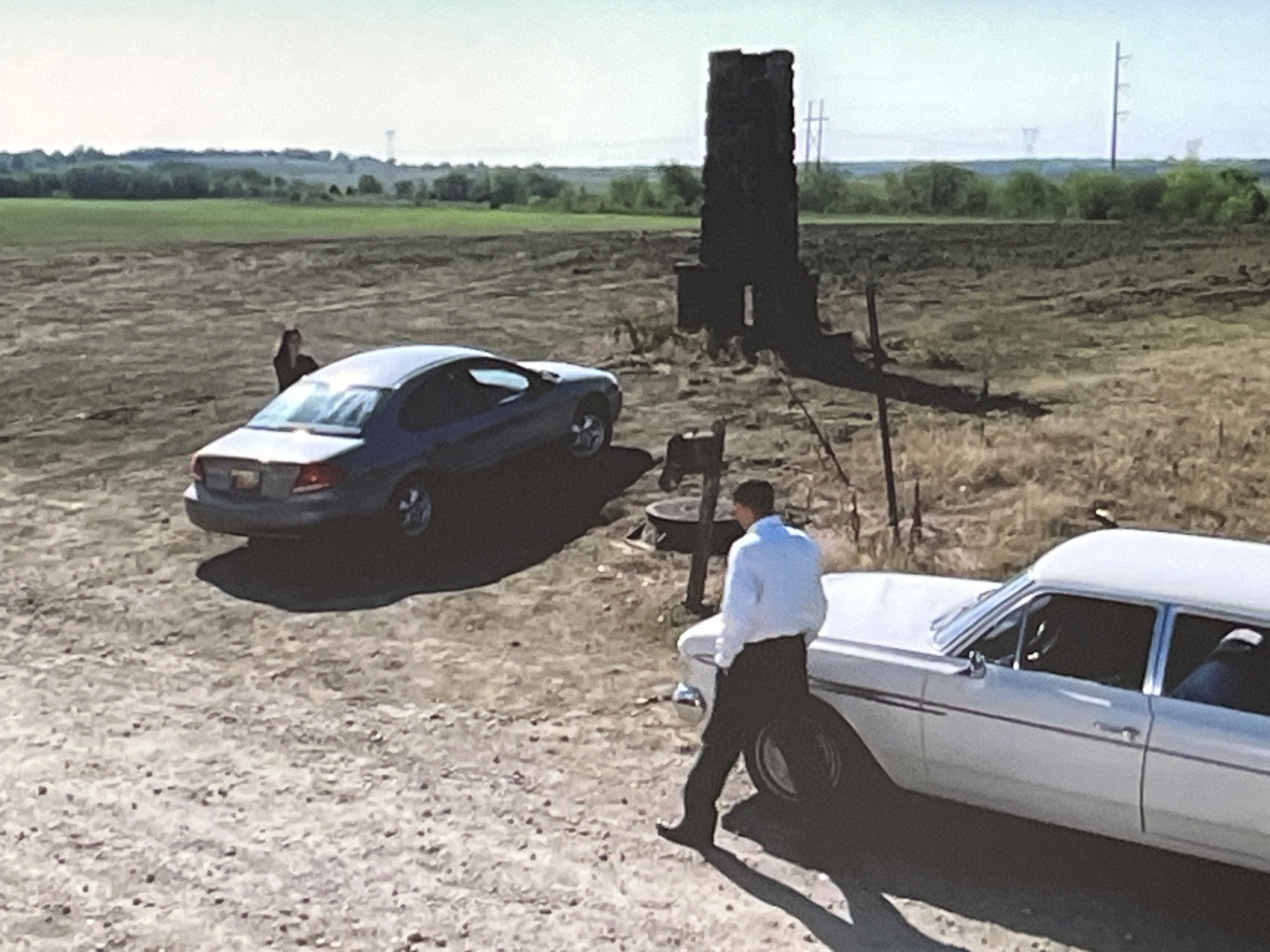 The image depicts an outdoor scene with two vehicles and two individuals. In the foreground, a white car, possibly a hearse, is partially visible on the right side of the frame. A person wearing a light blue shirt and dark pants is walking away from the car and towards the left. In the middle ground, a dark blue sedan is parked on a dirt or gravel surface. A person with dark hair is standing near the blue sedan. Behind the vehicles, a dark, rectangular structure, resembling a chimney or monument, stands in a field. The background shows a flat landscape with trees and a hazy sky. There are no identifiable landmarks or clues that would place this scene in Varna, Bulgaria.
