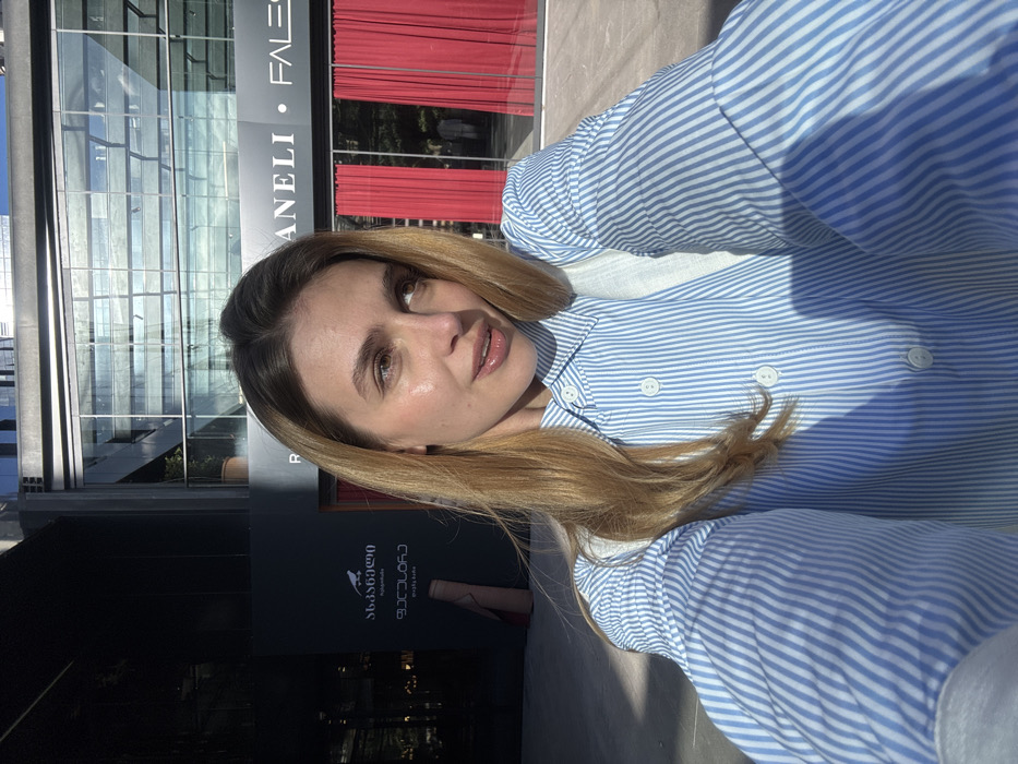 A young adult female is captured in a close-up selfie, standing outdoors in a commercial district of Tbilisi, Georgia, under bright sunlight. She has light-toned skin, long blonde hair, and is wearing a light blue and white vertical striped collared shirt. She looks towards her right, slightly upwards, with her head subtly tilted. Strong sunlight illuminates the right side of her face and hair, casting diagonal shadows across her attire. In the background to the left, a dark building facade is visible with white Georgian text, identifying "სანოტარო" (Notary), "ფასანაური" (Pasanuri), and "გოლეთი". To the right, a modern glass-fronted building features a sign with "ANELI • FALESI" and has its windows covered by prominent red vertical curtains. The architecture suggests a contemporary urban environment.