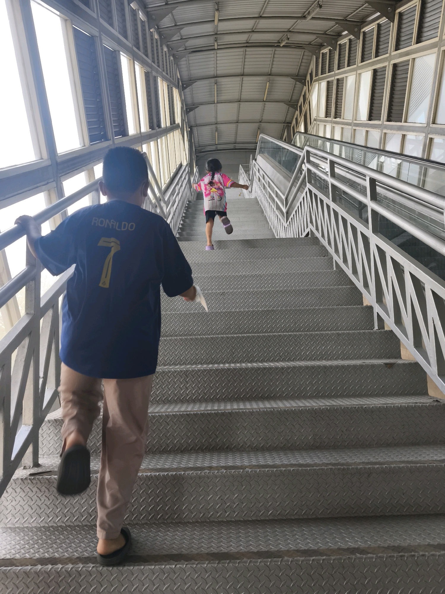 Two children are ascending a metal staircase. The child in the foreground, facing away from the camera, is wearing a dark blue t-shirt with "RONALDO 7" printed on the back and light brown pants. This child is wearing dark sandals. The second child, also facing away from the camera and further up the stairs, is wearing a pink and white top with a backpack and dark shorts, as well as pink and white socks with sandals. The staircase is made of diamond plate metal and has a silver metal railing. Large windows with venetian blinds are visible on the left and right sides of the staircase, suggesting an indoor or covered outdoor area. The ceiling is also metal. This scene is captured on an escalator, which is part of the sky bridge connecting the Palembang Icon mall to the Palembang Icon apartment complex, located in Palembang, Indonesia.