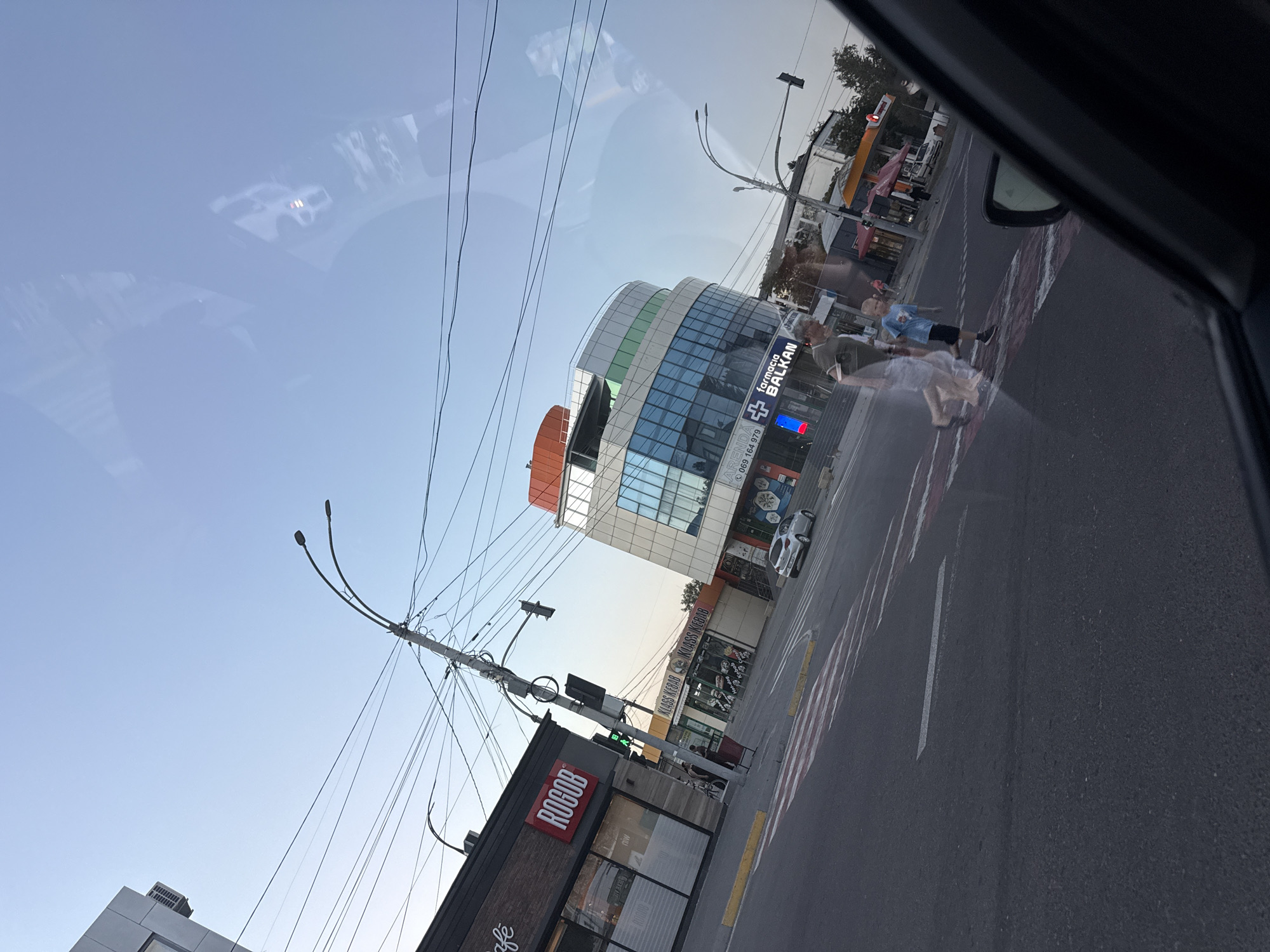 This is a street-level view of an intersection in Fundul Galbenei, Moldova, captured from inside a vehicle. The foreground shows the asphalt road with pedestrian crosswalk markings, including diagonal red and white stripes and parallel white lines. A silver car is visible in the middle ground, to the right of the intersection.
Several buildings line the street. To the left, a dark-colored building with a brick facade has signage that reads "ROGOB" in large red letters and "café" in smaller white text. Adjacent to it, a building with "NIW" visible on its windows. Further down the street, to the right, is a modern, multi-story building with a curved facade featuring blue glass windows and sections of white and orange cladding. A sign on this building reads "farmacia BALKAN". Another building with orange awnings and signage for "KLASS KEBAB" is also visible.
Several people are present. Two figures, one appearing to be an adult and a child, are crossing the street on the crosswalk in the center of the image. The adult is partially translucent due to a reflection or motion blur, while the child is clearly visible. Other pedestrians can be seen in the background near the buildings.
Overhead, a complex network of electrical wires and utility poles dominates the sky. Streetlights are also present. The sky is clear and light blue, indicating daytime. Reflections are visible on the surface from which the photo was taken, likely a car window, obscuring parts of the scene.