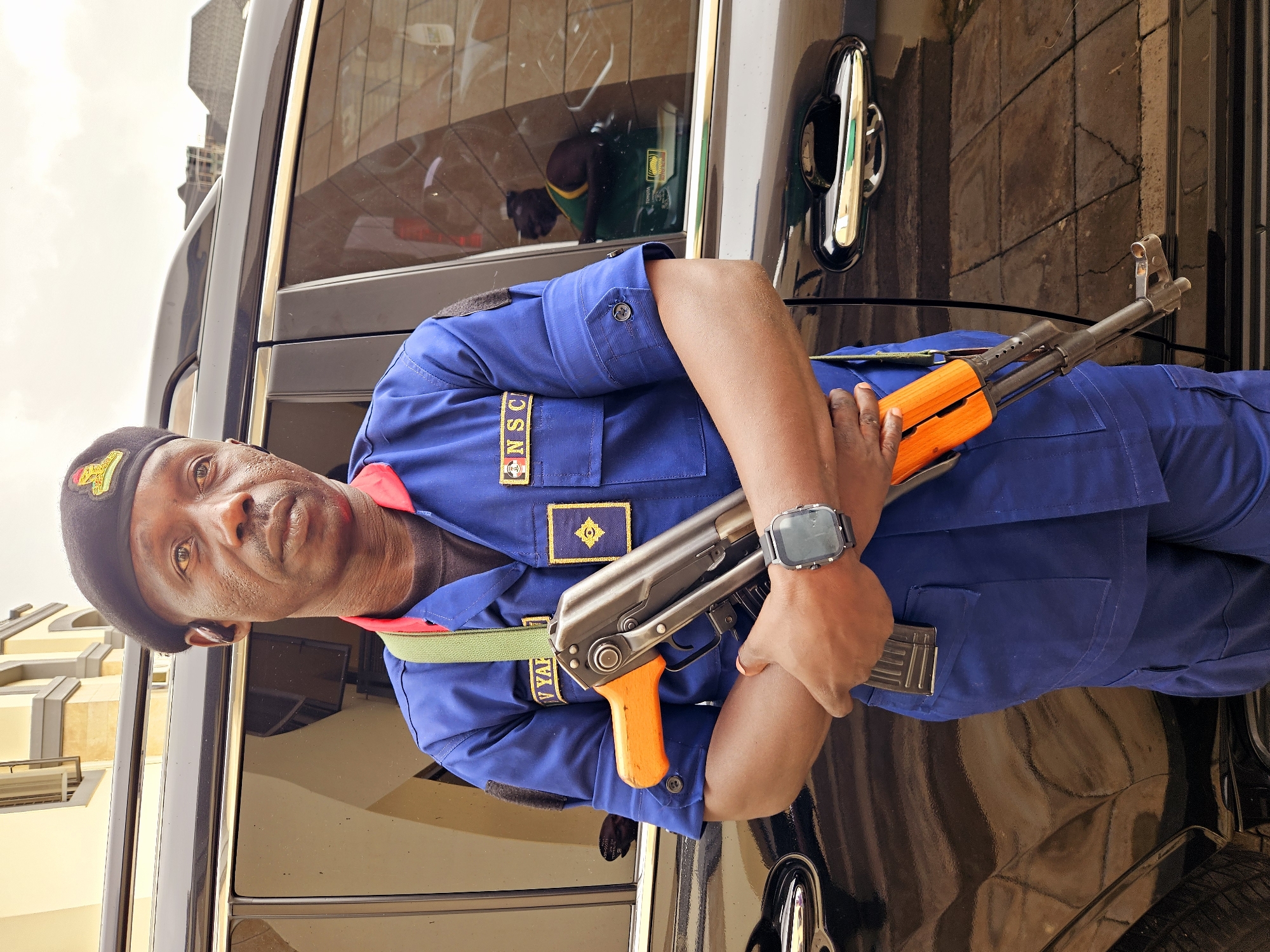 A Black male security guard in a blue uniform, black beret, and a green strap stands next to a black SUV in Guzape, Nigeria. The guard is holding an assault rifle with an orange stock and has a black smartwatch on his left wrist. The SUV's windows reflect a building and a person wearing a green shirt. The ground is paved with interlocking stones. The sky is cloudy.