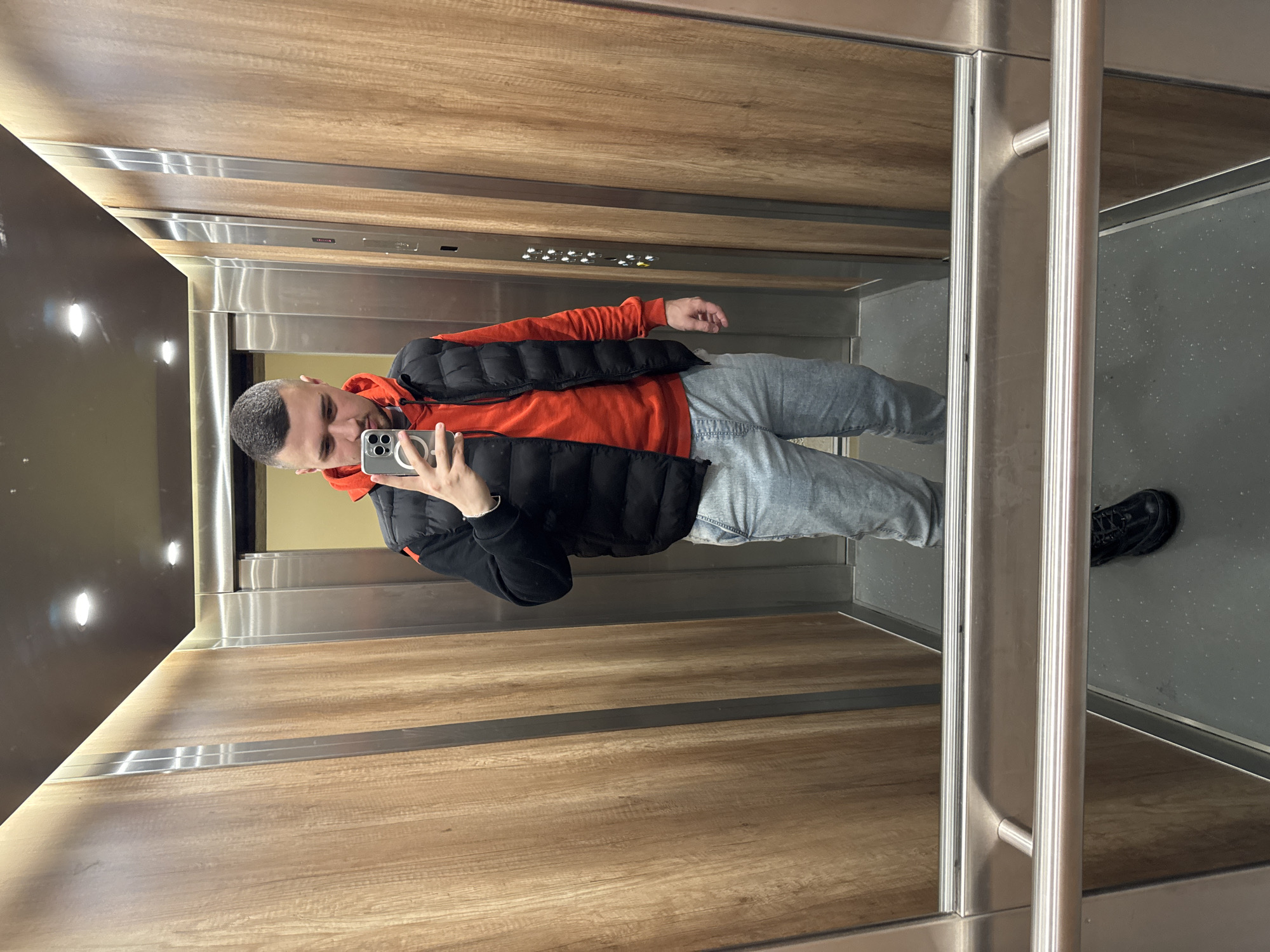 A man with short gray hair is standing in an elevator taking a selfie with a smartphone. He's wearing an orange hooded sweatshirt, a black puffer vest, and gray jeans. One black shoe is visible. The elevator's interior features wood-paneled walls with stainless steel accents, and the floor is gray. A set of elevator buttons are located above the man's head. The elevator door, framed by stainless steel, is to his right. The location is not identifiable as Sofia, Bulgaria.