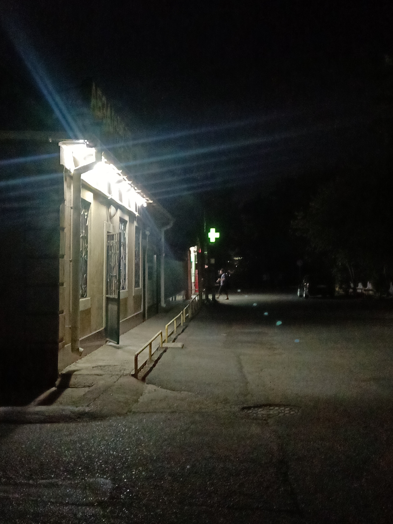 An outdoor scene at night in Shymkent, Kazakhstan, depicts the facade of a building illuminated by overhead lights, casting bright reflections on the wet pavement. The building, appearing to be a pharmacy with a bright green cross sign prominently displayed, has barred windows and an open door. A yellow metal railing runs along the sidewalk in front of the building. A person is visible walking away from the building towards the right side of the frame, and in the background, a dark vehicle is parked. The environment is dark with ambient lighting from the building and the green cross, with some light streaks across the sky, possibly from a light source not visible in the frame.
