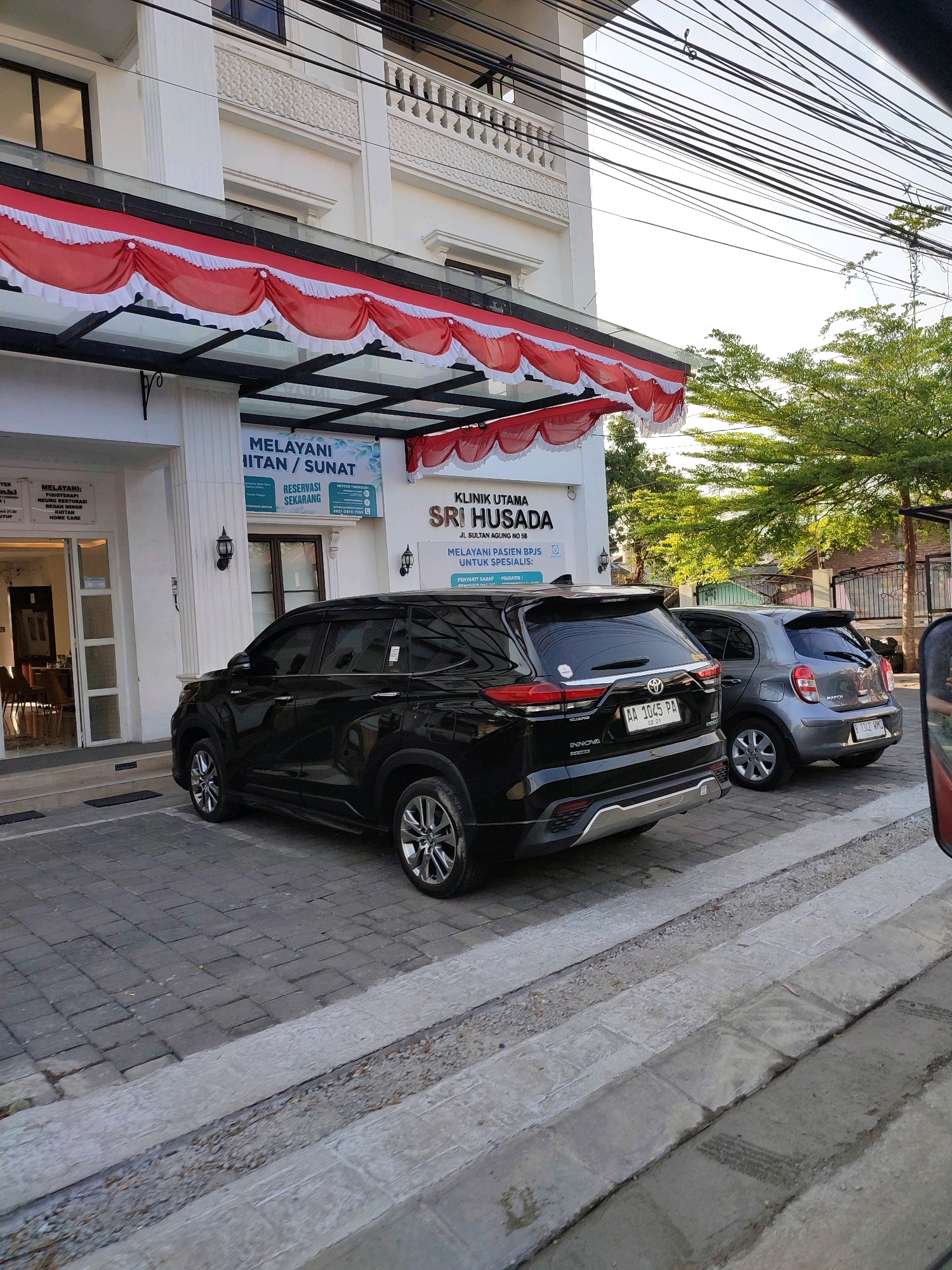 The image shows a clinic named "KLINIK UTAMA SRI HUSADA" located in Magelang, Indonesia. The clinic has a white facade with red and white decorations above the entrance. Two cars are parked in front of the clinic: a black SUV and a gray hatchback. The black SUV is a Toyota Innova with license plate AA 1045 PA. The gray hatchback is a Nissan March with license plate B 1042 MHI. The area is paved with interlocking bricks and there is a drainage ditch in the foreground. The sky is clear and the sun is setting, casting a warm light on the scene. The overall impression is of a clean and well-maintained clinic on a quiet street.