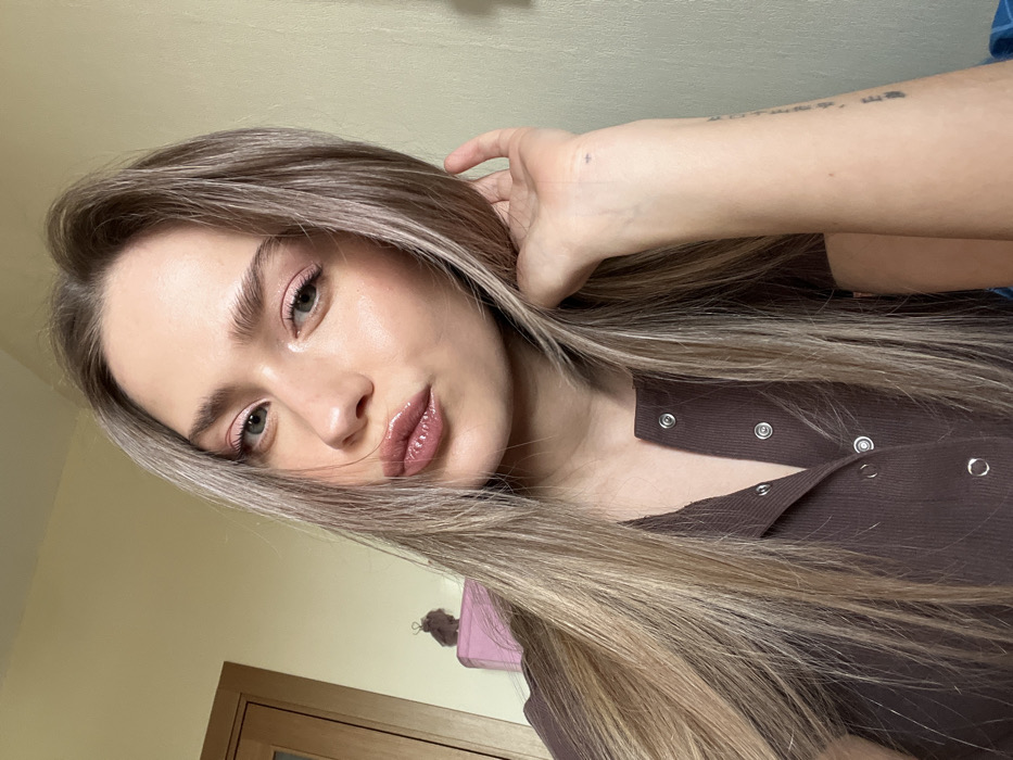 A young adult female is featured in a close-up indoor photograph, looking directly at the camera. She has long, multi-toned light brown hair with blonde highlights, light-colored eyes, pink eyeshadow, and glossy lips with a slight pucker. She is wearing a dark brown, ribbed short-sleeved top with several unfastened snap buttons down the front. Her left hand is positioned near her left ear, with fingers touching her hair. A small dark mark is visible on her left wrist, and faint, vertical text, possibly a tattoo, is present on her outer left forearm. The background is blurred, revealing a plain, light-colored wall. To the left, a wooden door and its frame are partially visible, above a pink rectangular object on which a small, dark brown, textured item rests. The scene takes place within a generic interior setting. There are no visual elements within the image that enable the identification of a specific location in Saint Petersburg, Russia.