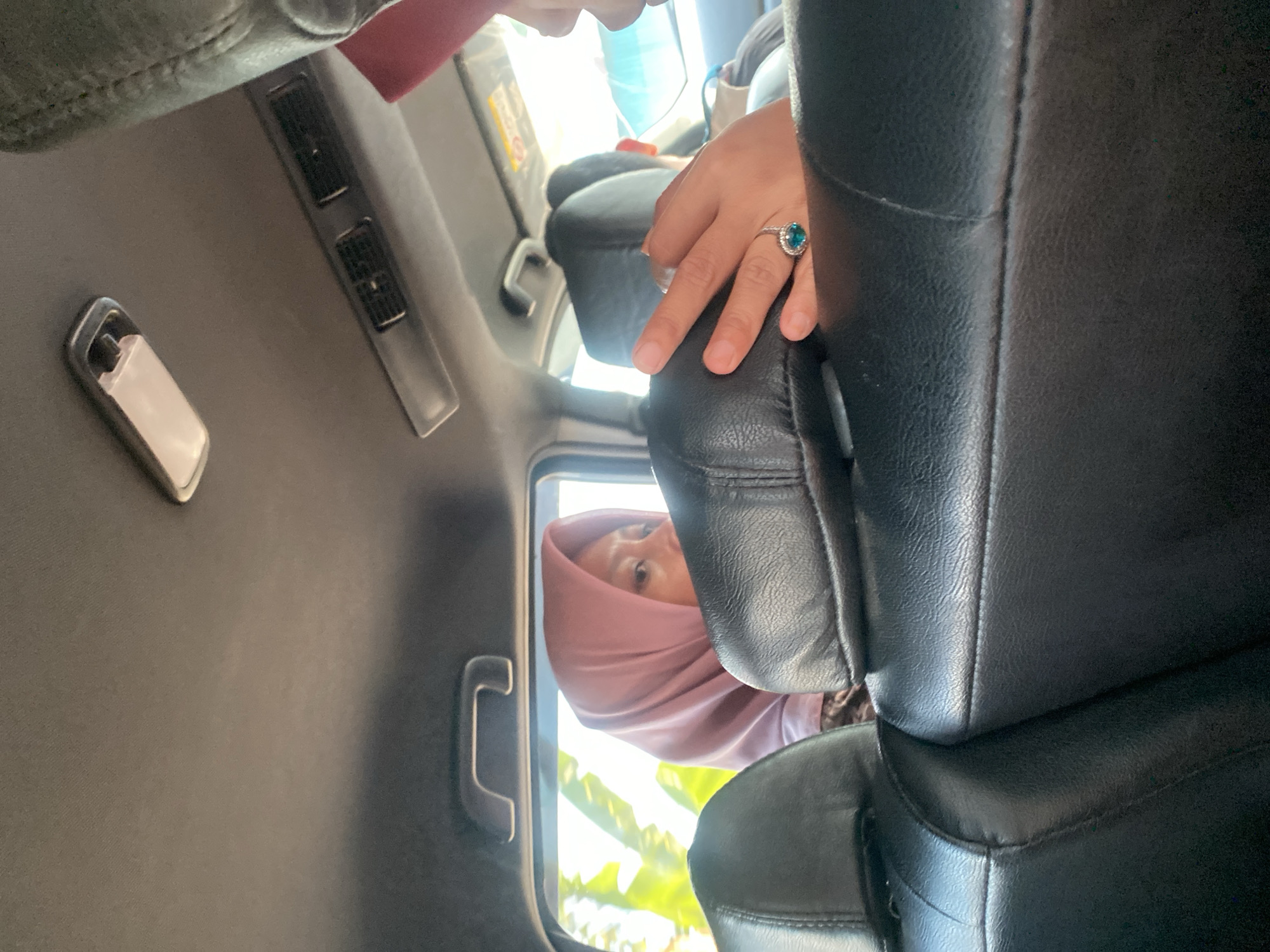 The image shows the interior of a car, likely in Antasan Senor Ilir, Indonesia, based on the provided context. In the foreground, black leather car seats are visible. Behind the seats, a woman wearing a pink hijab is looking out the window. Her face is partially obscured by the car's headrest. Her right hand, adorned with a ring featuring a turquoise gemstone, rests on the headrest. The background reveals a bright outdoor scene with green foliage, suggesting a daytime setting. The car's interior also shows a ceiling light and air conditioning vents.