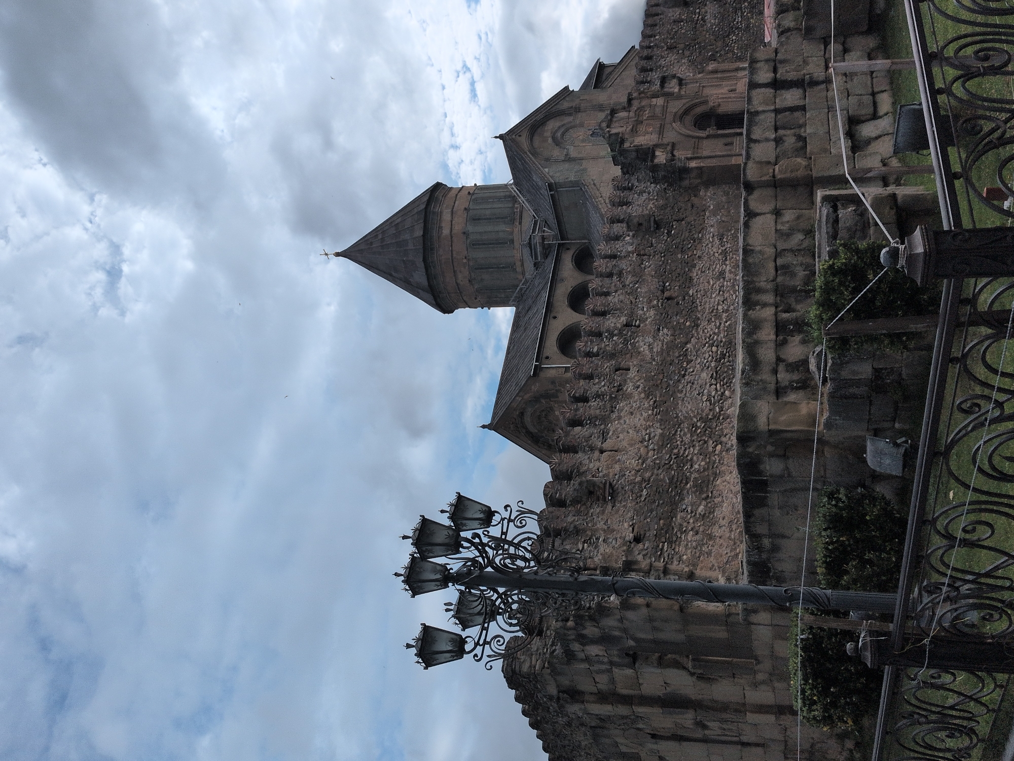 The image depicts the Svetitskhoveli Cathedral in Mtskheta, Georgia, a UNESCO World Heritage Site. The cathedral, a prominent example of medieval Georgian architecture, is constructed of stone and features a large central dome topped with a cross. A fortified stone wall with crenellations surrounds the cathedral complex, contributing to its imposing appearance. In the foreground, a decorative lamppost with multiple lamps stands tall, casting shadows on the weathered stone. Greenery, including bushes and grass, is visible within the walled area and beyond a black ornamental fence. The sky above is overcast with dramatic clouds, suggesting a potentially moody or stormy atmosphere. The scene is static and lacks any visible human presence or activity.