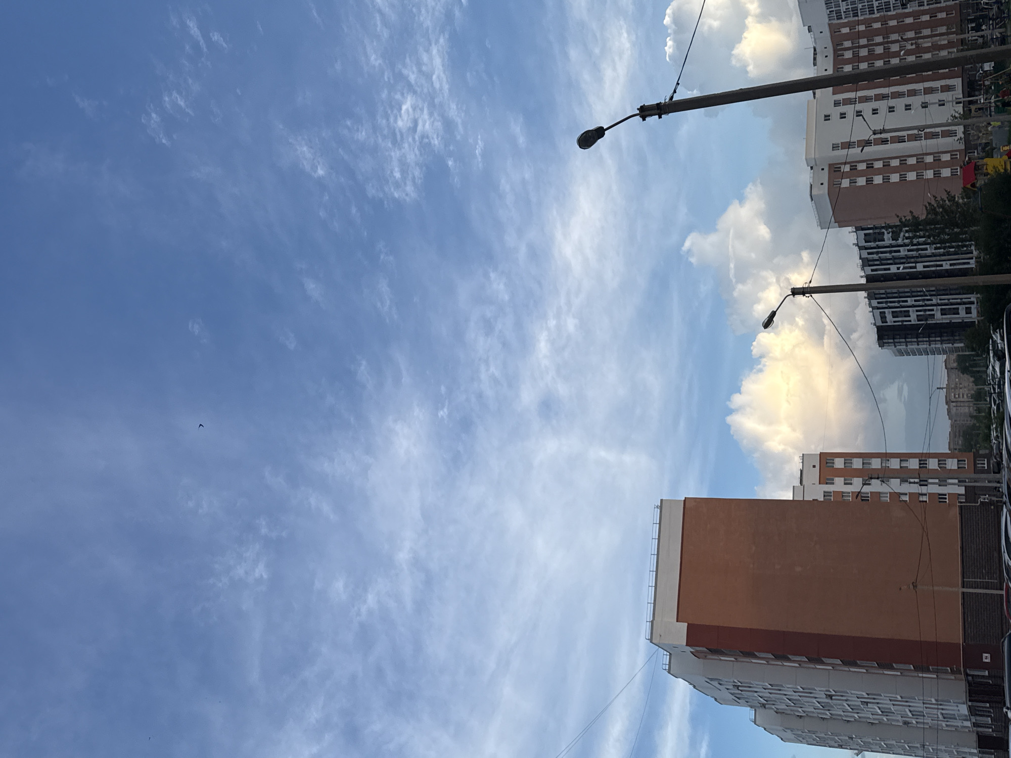The image shows a bright blue sky with wispy white clouds. In the foreground, there are several tall apartment buildings. The building on the left is white with a red and orange section. The building in the center is mostly white with orange accents. The building on the right is white with orange windows. A street lamp is visible on the right side of the image. The scene appears to be in a residential area of a city. There are no people clearly visible in the image.