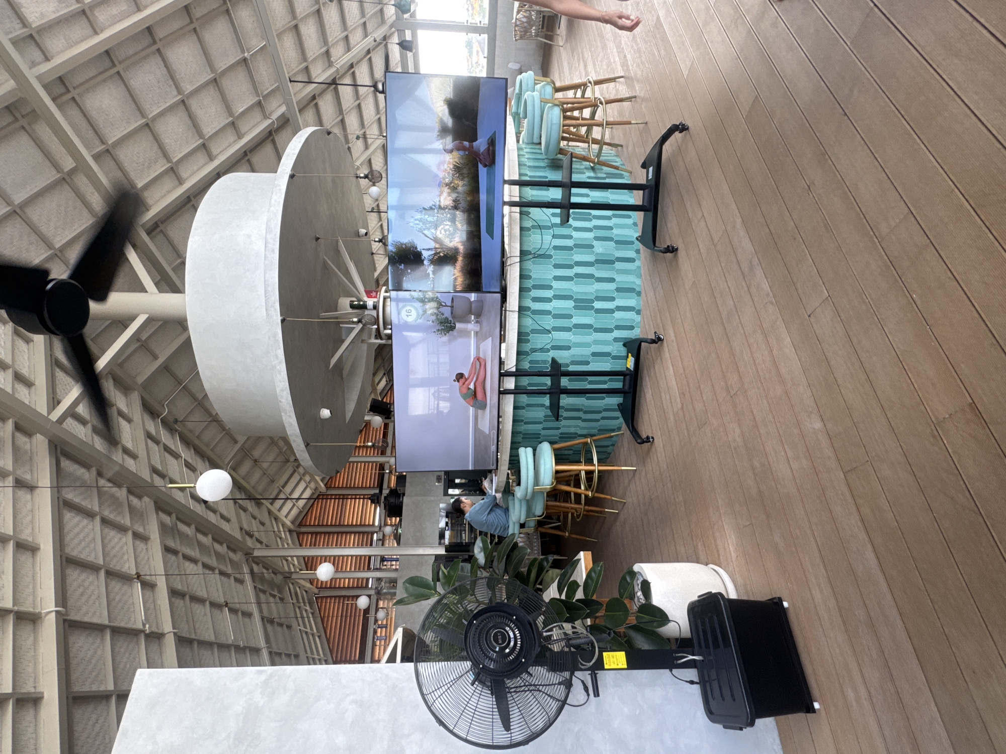 The image depicts the interior of a restaurant or cafe located in North Kuta, Indonesia. The space features a light-colored, slatted ceiling with exposed beams, and large industrial-style fans suspended from above. Several spherical pendant lights hang from the ceiling, contributing to the ambient lighting.
In the foreground, a large, black industrial fan stands to the left, positioned near a potted plant and a black plastic bin. The floor is made of light brown wooden planks. A bar area with teal-tiled mosaic patterns forms the central focus. Behind the bar, two large flat-screen televisions are mounted on black stands, displaying what appears to be a yoga or fitness class. The screens show individuals performing yoga poses in serene outdoor settings.
Several light blue bar stools with golden legs are arranged around the bar. A person, dressed in a blue shirt, is seated at the bar, facing the televisions and appearing to be engaged with the content on the screens. In the background, large windows allow natural light to enter, and a glimpse of an outdoor seating area with wicker furniture can be seen. The overall atmosphere suggests a modern, relaxed, and well-lit establishment.