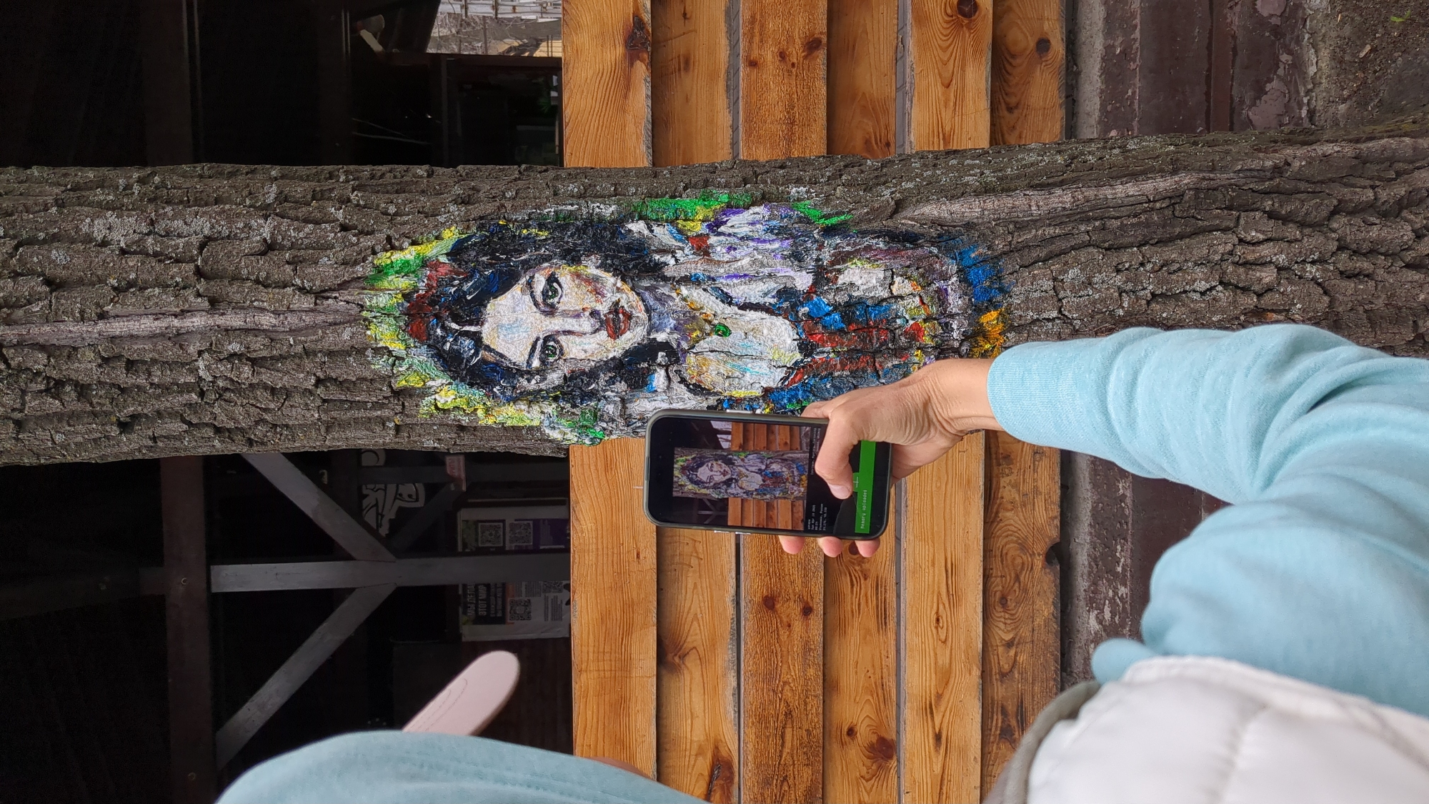 A person is holding a cell phone to take a photo of a mural in Ulyanovsk, Russia. The mural, depicting a woman's face, is painted on a fallen tree trunk. The trunk is situated in front of a wooden wall. The person holding the phone is wearing a light blue sweater and a white padded jacket. The phone screen displays a picture of the mural. The background includes a wooden structure and a glimpse into a dimly lit interior.