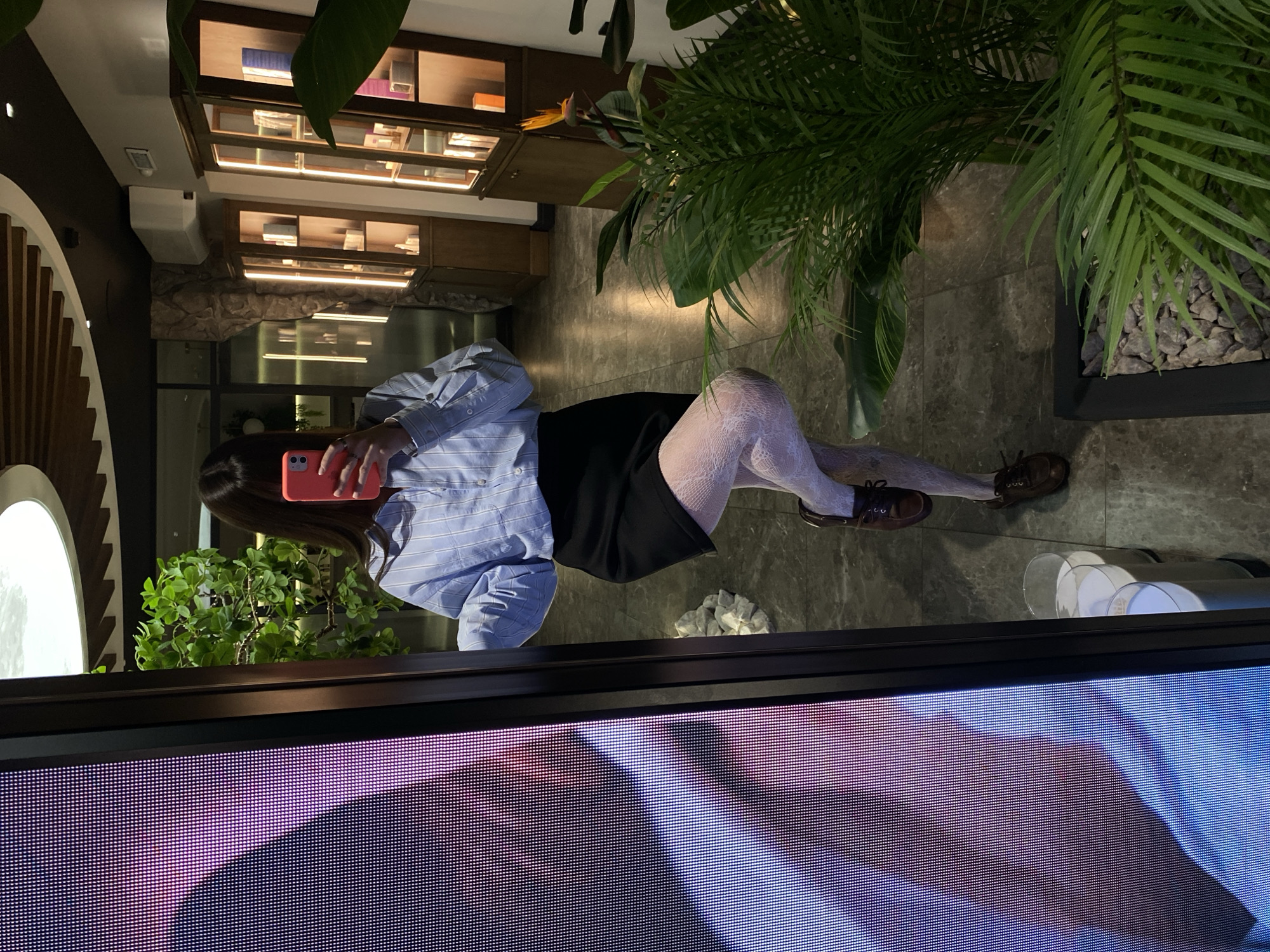 A young woman is photographed from a low angle, seemingly reflected in a large screen or monitor. The woman is holding a pink smartphone in front of her face, obscuring it. She is wearing a light blue striped, button-down shirt, a black skirt, and white patterned tights. Her feet are clad in brown lace-up shoes.
The background includes interior design elements such as shelving with items, a round decorative element, a potted plant, and what appears to be a marble-like floor.
The lower portion of the frame is dominated by the screen reflecting the woman, displaying a pattern of red and blue pixels.
The location is likely indoors, possibly a store or cafe based on the interior design. It is not possible to verify if this is Votkinsk, Russia.