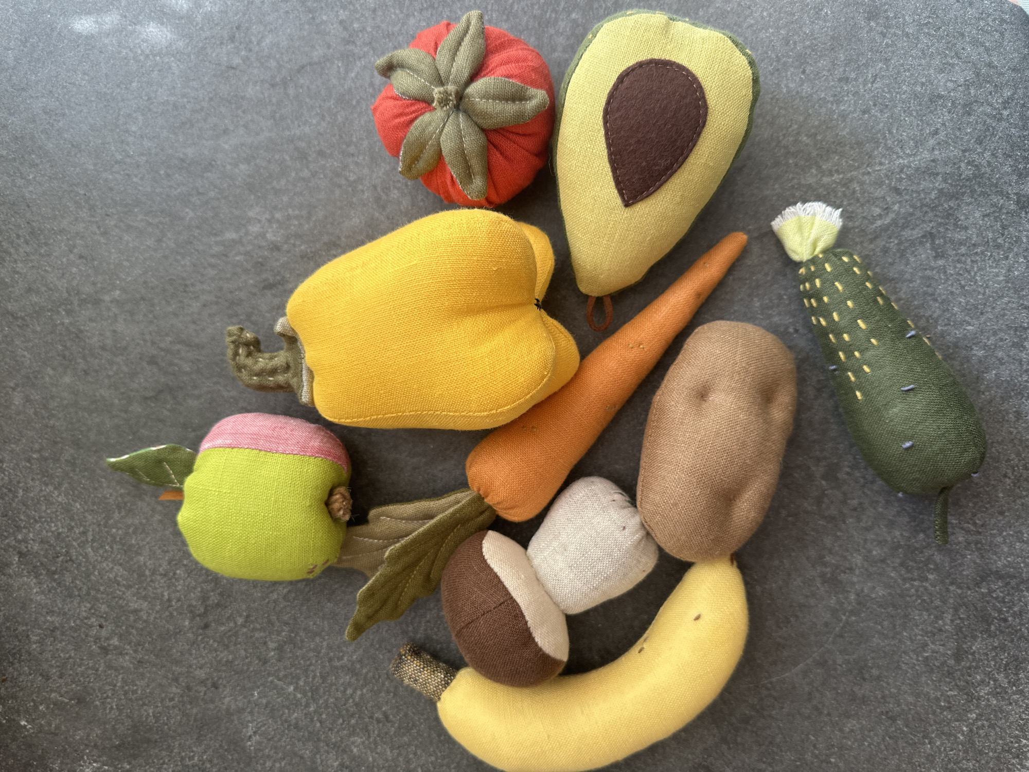 This image displays a collection of handmade plush toys resembling various fruits and vegetables arranged on a dark gray, textured surface. The toys include an apple, a yellow bell pepper, a tomato, a banana, a mushroom, a potato, a carrot, an avocado half, and a cucumber. The background appears to be a flat, possibly stone or concrete surface, with some subtle variations in texture and color. There are no people or actions depicted in the image. The lighting suggests an indoor setting, with soft shadows cast by the objects. The exact location in Chornomorske, Ukraine, is not discernible from the visual information provided.