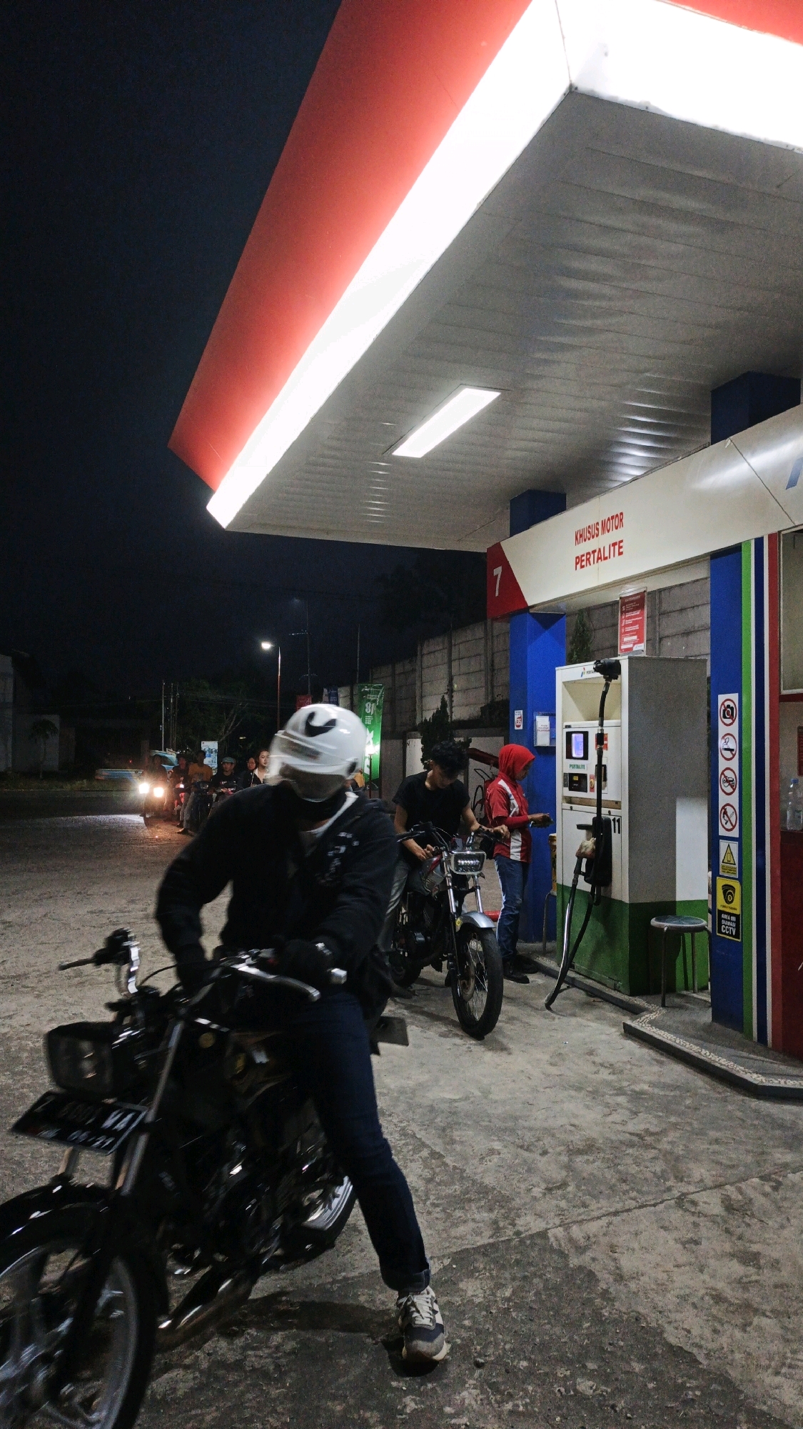 The image depicts a nighttime scene at a gas station, likely in Sukaresik, Indonesia. The foreground is dominated by a person wearing a black jacket and blue jeans, sitting on a black motorcycle. They are also wearing a white helmet with a black Nike logo. The motorcycle has a license plate that reads "Z 6001 WA". Behind this person, another motorcycle is visible with two individuals. One is wearing a black t-shirt and the other is wearing a red hooded jacket and jeans. They are standing near a gas pump. The gas station has a red and white awning with fluorescent lighting. A sign above the pump reads "KHUSUS MOTOR PERTALITE". In the background, other motorcycles and a dimly lit street are visible. The overall environment suggests a casual gathering of motorcyclists at a gas station after dark.