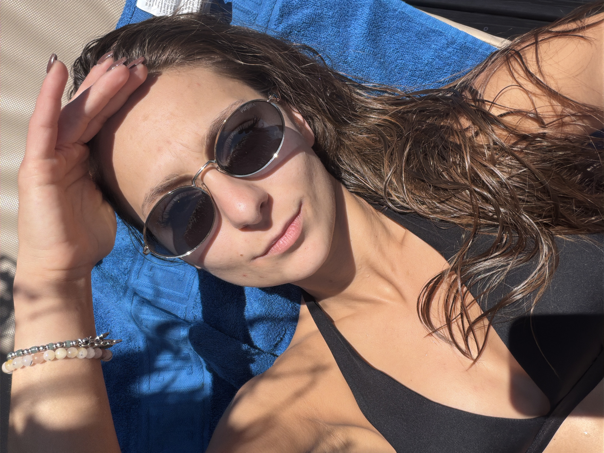 A young woman with long, wet, brown hair and light skin is lying on a blue towel outdoors, likely on a lounge chair given the background. She is wearing a black bikini top and sunglasses with thin, silver frames. Her right hand is raised, shielding her eyes from the sun, and she is wearing a bracelet on her left wrist composed of light-colored beads and a silver charm. The lighting suggests it is a sunny day, with strong shadows cast on her body and the towel, indicative of overhead light sources and possibly tree foliage. There is a faint shadow on her forehead. The scene does not provide any specific visual cues to determine the location within the city of Plovdiv, Bulgaria.