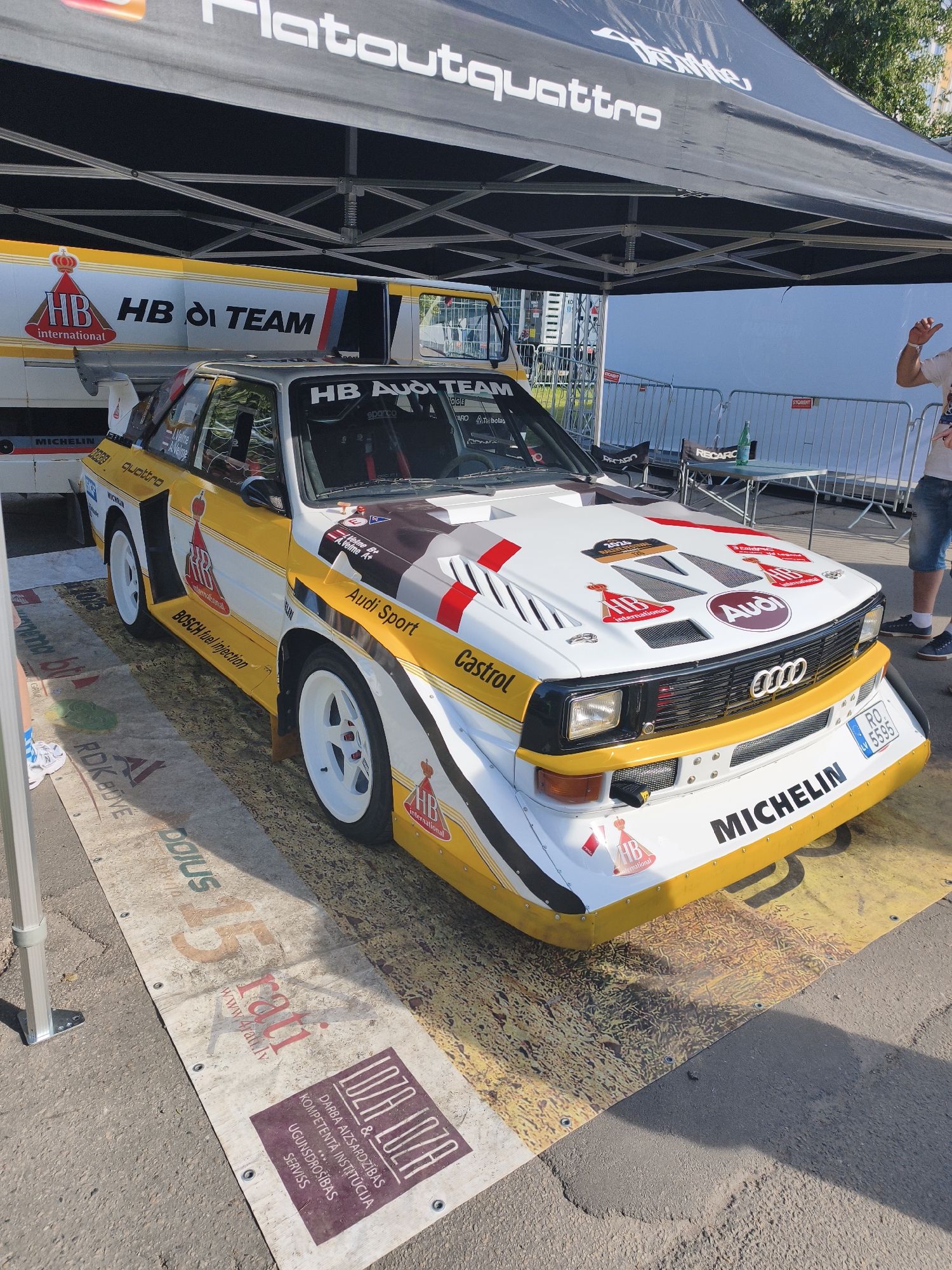 The image shows a rally car, an Audi Sport Quattro, parked under a black canopy tent with "Flatoutquattro" branding. The car is white with yellow and black racing stripes and features numerous sponsor decals, including "HB HB International," "Audi Sport," "Castrol," and "Michelin." The license plate reads "RO 5595." The car is positioned on a wooden platform covered with a printed mat that has various logos and text, including "15," "rati," and "LOZA LOZA & DARBA AIZSARDZĪBAS KOMPETENTĀ INSTITŪCIJA UGUNSGRĒKS SERVISS". In the background, there is a white wall with some metal fencing and a small table with chairs, one of which has a "RECARO" logo. A person in a white t-shirt and denim shorts is partially visible on the right side of the frame, gesturing with their hand. Another person's legs, wearing white sneakers with blue stripes, are visible on the left. The scene appears to be an outdoor event, possibly a car show or rally exhibition, taking place on a sunny day. The presence of the "rati" logo and the Latvian text on the mat suggests the location is Riga, Latvia, specifically at the 15th-anniversary celebration of Rati, a Latvian automotive club, which took place on June 10, 2023, at the Biķernieki circuit in Riga.
