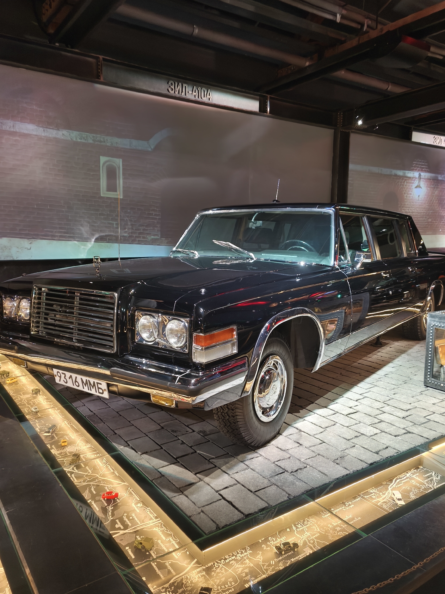 A black ZIL-4104 limousine is displayed in a museum, possibly in Moscow, Russia, given the Cyrillic signage reading "ЗИЛ-4104" above the vehicle. The limousine is positioned on a cobblestone-like floor and is viewed from a slightly elevated, forward-angled perspective. The vehicle has a long wheelbase, chrome accents, and distinctive round headlights. In the foreground, a display case with a map and miniature vehicles is illuminated. The background features a brick wall projection with an arched window and overhead industrial-style piping and lighting. There are no people visible in the image.