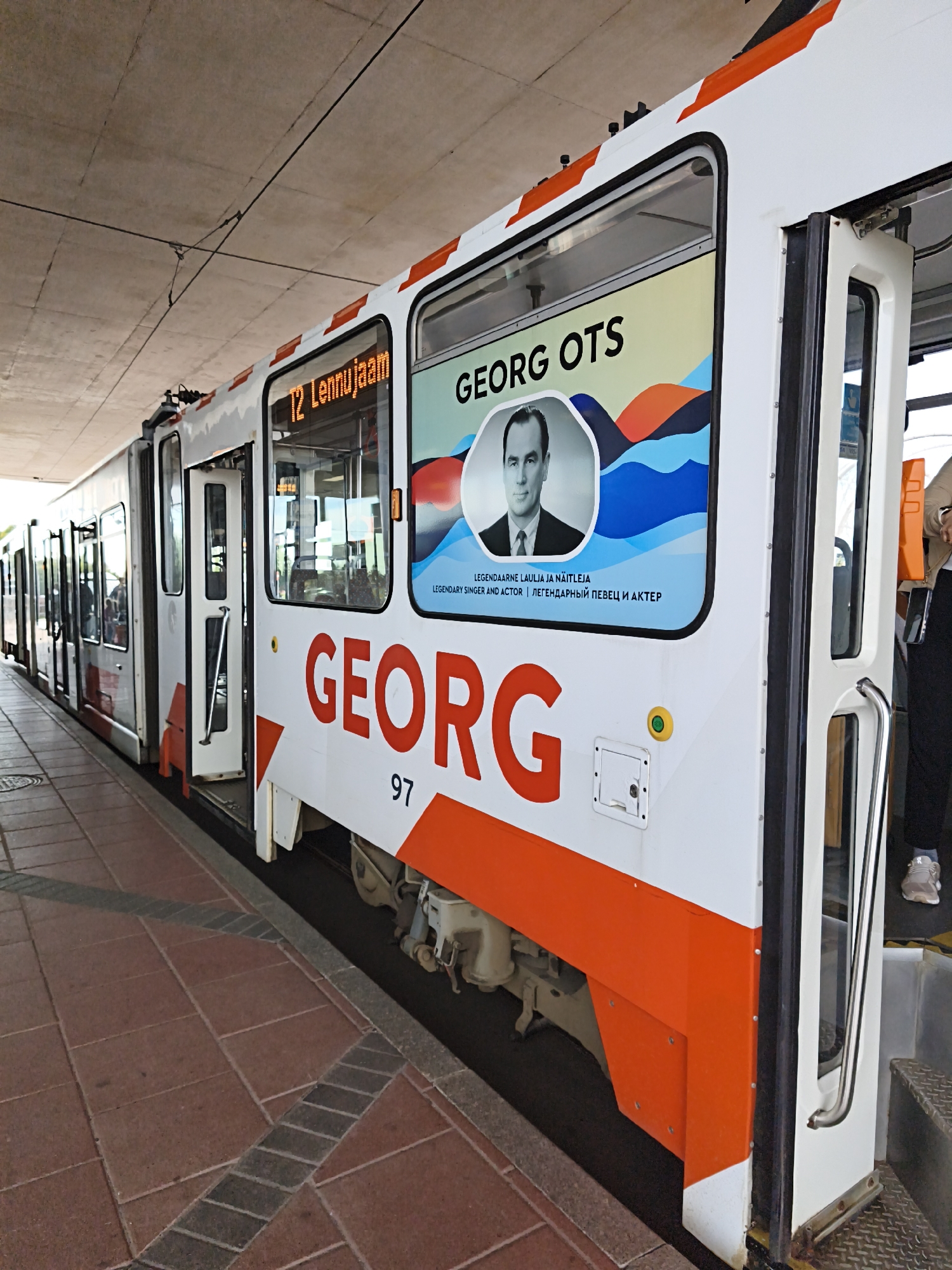 A tram is parked at a station with red tiled flooring and a concrete ceiling overhead. The tram is white with orange accents and features a portrait of Georg Ots on its side, along with the text "GEORG OTS" and "LEGENDARY SINGER AND ACTOR". The number "97" is also visible on the tram. The tram doors are open, and passengers can be seen inside. The scene appears to be in Tallinn, Estonia, given the prominent display of Georg Ots, a renowned Estonian singer.