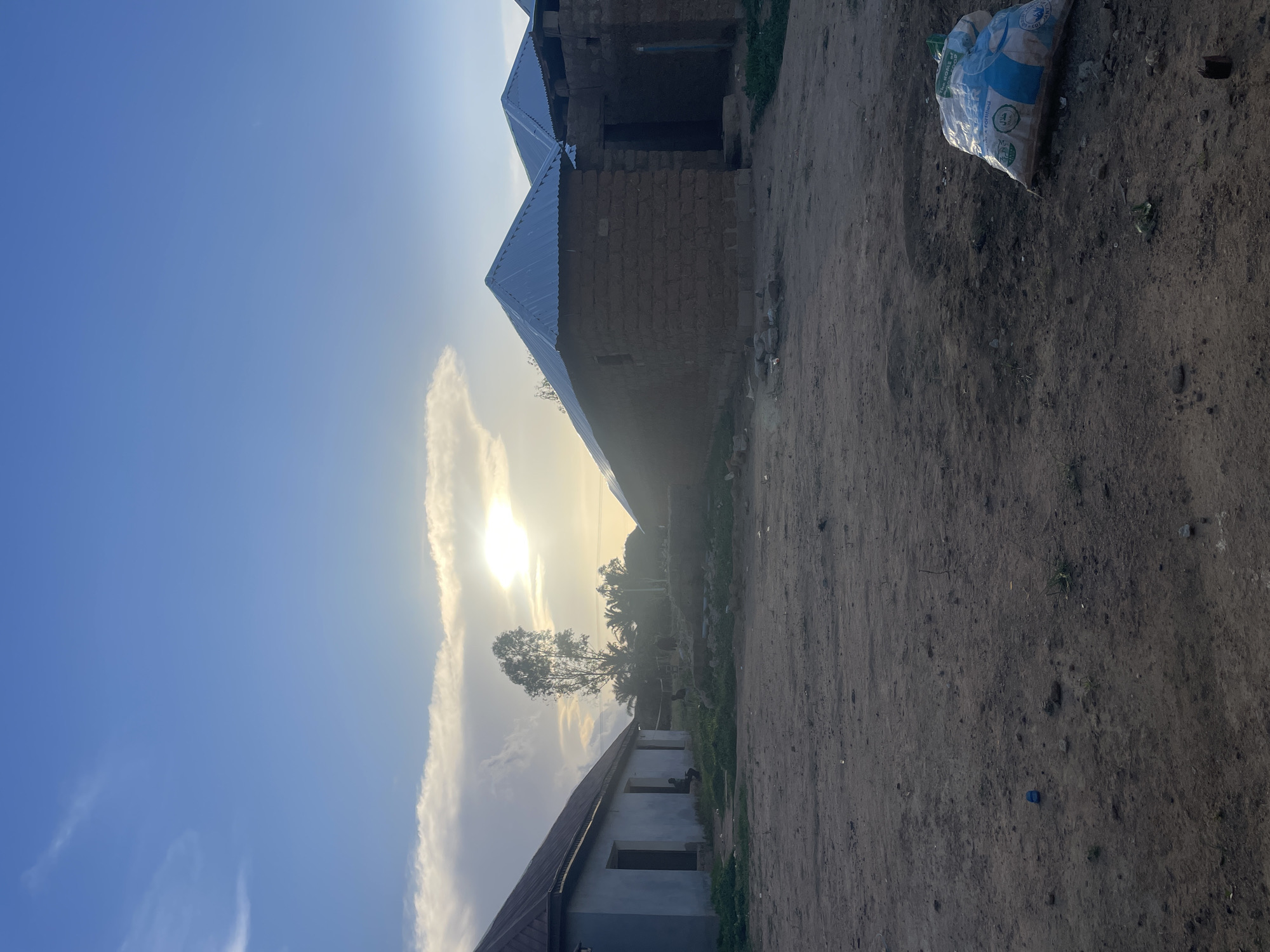 The image depicts a rural scene, presumably in Durimi, Nigeria. A dirt path runs through the right side of the frame. A partly full bag sits by the path's edge. Buildings with corrugated metal roofs are scattered. One building appears to be under construction with unfinished brick walls. In the background, trees are partially obscured by a bright sun and cloud formation. A lower building with open rectangular window spaces sits beside the path.