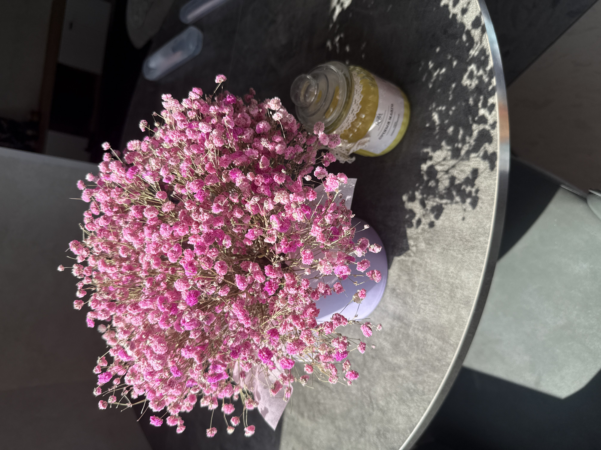 A high-angle shot reveals a close-up of a bouquet of pink baby's breath flowers in a light purple vase on a gray, round table. Next to the flowers sits a glass jar with a yellow substance inside and a white lace ribbon wrapped around the top. Sunlight casts a shadow of leaves on the table's surface. The setting is likely indoors due to the controlled lighting and arrangement of objects. The location is not apparent from the image.