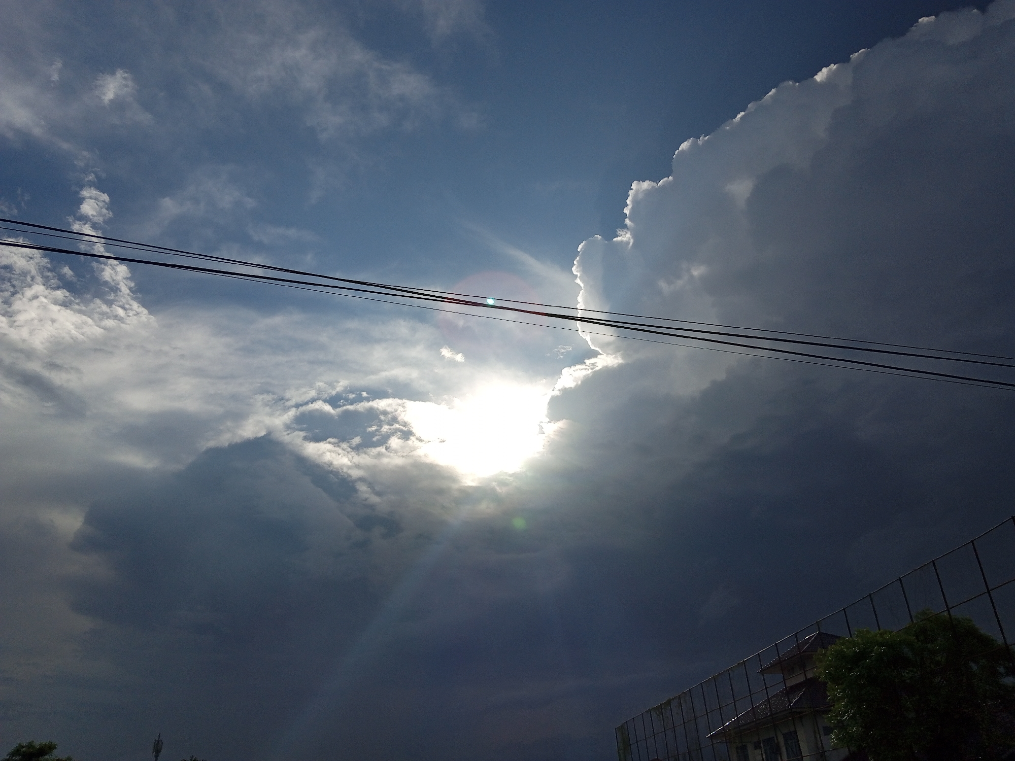 The image, taken in Uteunreutoh, Indonesia, presents a cloudy sky with a bright sun obscured by clouds. Two parallel black electrical wires stretch across the upper part of the frame. The cloud cover varies from lighter white wisps to dark, ominous storm clouds. In the lower right corner, a building is partially visible behind a chain-link fence, with a green tree obscuring some of the building's facade. Some trees are visible on the lower left. An antenna is visible to the left of the trees, at the bottom center.